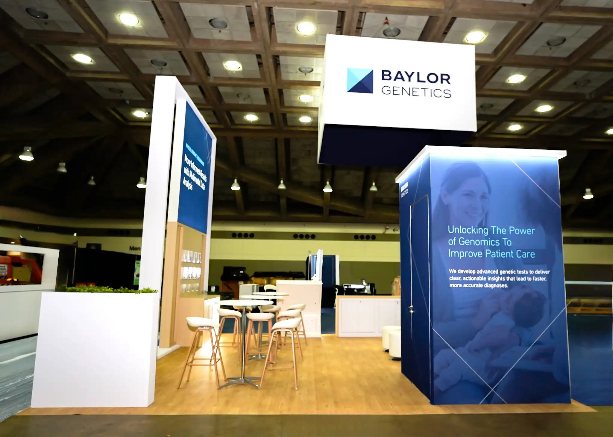 Side angle of Baylor Genetics booth showing large Unlocking The Power of Genomics photo mural panel, beverage hospitality station, round bar-height tables, live plants, DNA helix pattern graphics, and warm ambient lighting creating an inviting scientific environment