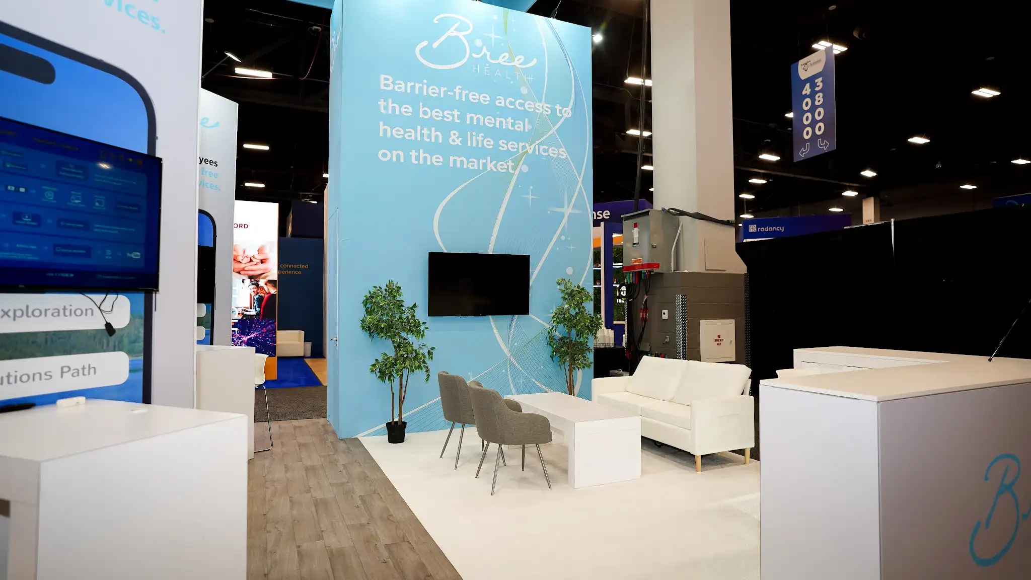 Interior ground-level view of the Bree Health 20x30 ft island booth at HR Tech 2025 looking toward the primary backwall showing the tall full-height backwall wrapped in vivid light sky blue graphic with flowing abstract white swooping ribbon line patterns, Bree Health cursive logo at the top, tagline Barrier-free access to the best mental health and life services on the market, centrally wall-mounted flat-screen TV, two tall potted green trees in black pots flanking the TV adding natural welcoming element, white modern couch sofa on the right side, two round-backed gray upholstered accent chairs in the center, white rectangular low coffee table, warm wide-plank wood-look vinyl flooring, left side showing tall white panel displays with smartphone mockup graphics of the Bree Health app with Exploration and Solutions Path text visible, white counter kiosk structure at far right with cursive Bree logo in teal, Radancy booth visible in background, booth number signage 43800 and 40000 visible at top