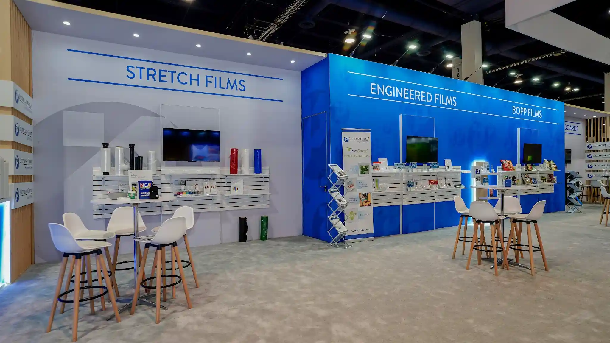 Close interior view of Inteplast Group booth showing Stretch Films section on white background and Engineered Films section on bright blue background, with white slatwall product displays holding film rolls in multiple colors, bar-height tables with white molded seats on wooden legs, and multiple flat-screen monitors displaying product content