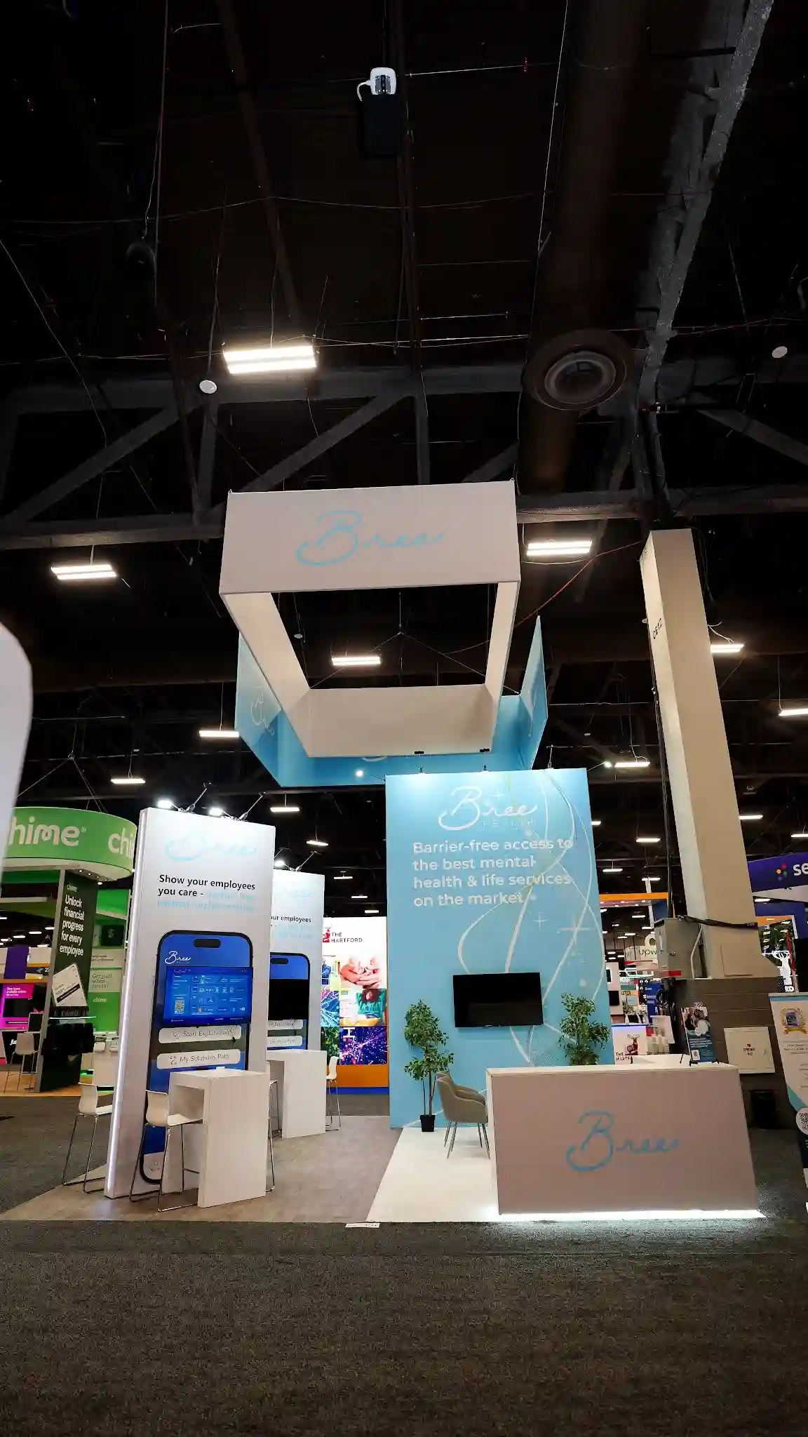 Front exterior view of the complete Bree Health booth at HR Tech 2025 showing both the left kiosk tower section with Bree logo and Show your employees you care text with large phone graphic of the Bree app and TV monitor, and the right backwall lounge section with light blue backwall displaying Bree Health logo and tagline with wall-mounted TV and potted green tree and gray accent chair, white reception counter at bottom right with cursive Bree logo in teal and LED accent strip, overhead hanging sign structure in white and sky blue with Bree Health branding suspended by cables, neighboring Chime green booth on left and Sense blue booth on right visible in background, Mandalay Bay convention center dark ceiling