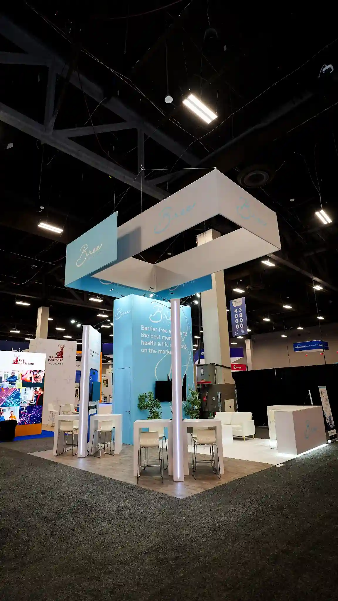 Wider diagonal exterior view of the Bree Health 20x30 ft island booth at HR Tech 2025 showing the full booth layout with large white-dominant overhead hanging sign with cursive Bree logo in teal on white face and adjacent light blue panel suspended by thin cables with built-in underside lighting, tall central backwall in light sky blue prominently displaying Bree logo and tagline Barrier-free access to the best mental health and life services on the market, interior seating area with white high-top bar tables and light wood natural tone bar stools in cluster arrangement, white modern armchairs sofas visible toward the right, potted plant near backwall, warm wood-look laminate flooring, two tall white illuminated column-like pylon structures flanking interior adding vertical accent lighting, white reception demo counter labeled with cursive Bree logo in teal on the right, The Hartford insurance booth with stag logo visible in background to the left, Radancy booth faintly visible, aisle number sign 43800 40000 in upper right, Mandalay Bay convention center dark ceiling