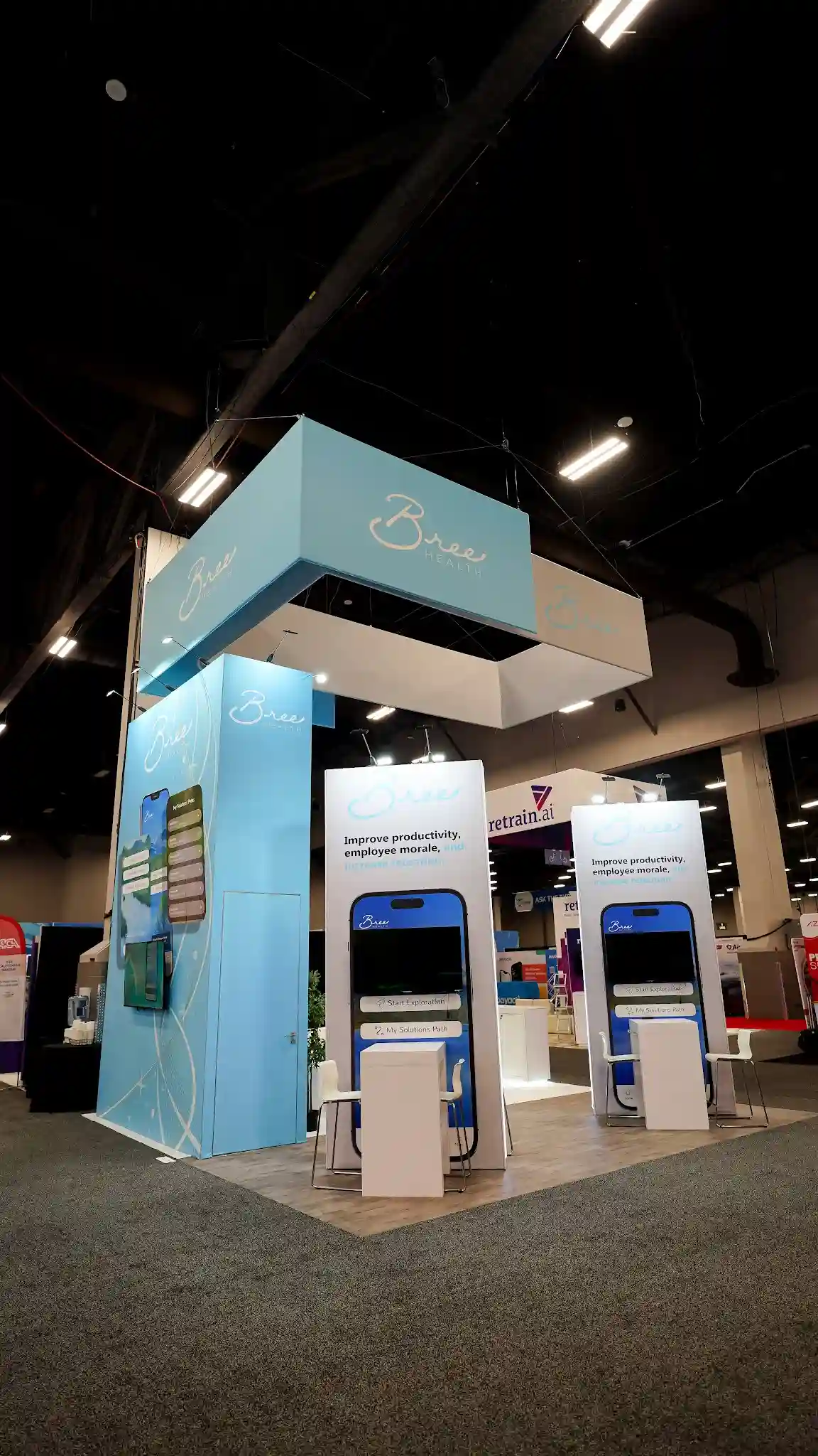 Front corner view of the Bree Health booth showing two identical tall white interactive kiosk stations with large phone silhouette graphics in navy blue displaying the Bree app and Improve productivity employee morale messaging, asymmetric overhead hanging sign in white and sky blue with Bree Health branding, central tower visible behind kiosks, retrain.ai booth in background