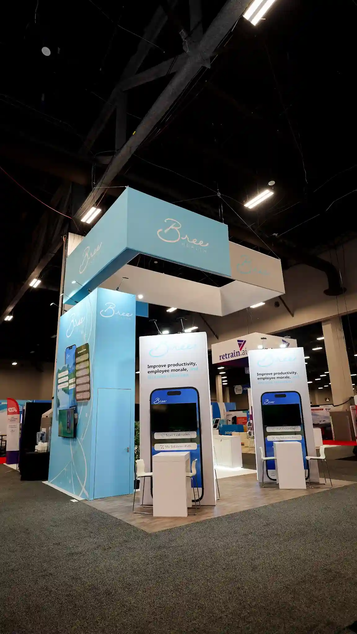 Similar front angle of the Bree Health booth showing both interactive kiosk stations with phone graphics and Improve productivity employee morale text, multi-panel overhead hanging sign with intersecting white and sky blue elements, main tower on left side with Bree logo and app graphics, white pedestals and bar chairs, convention hall setting