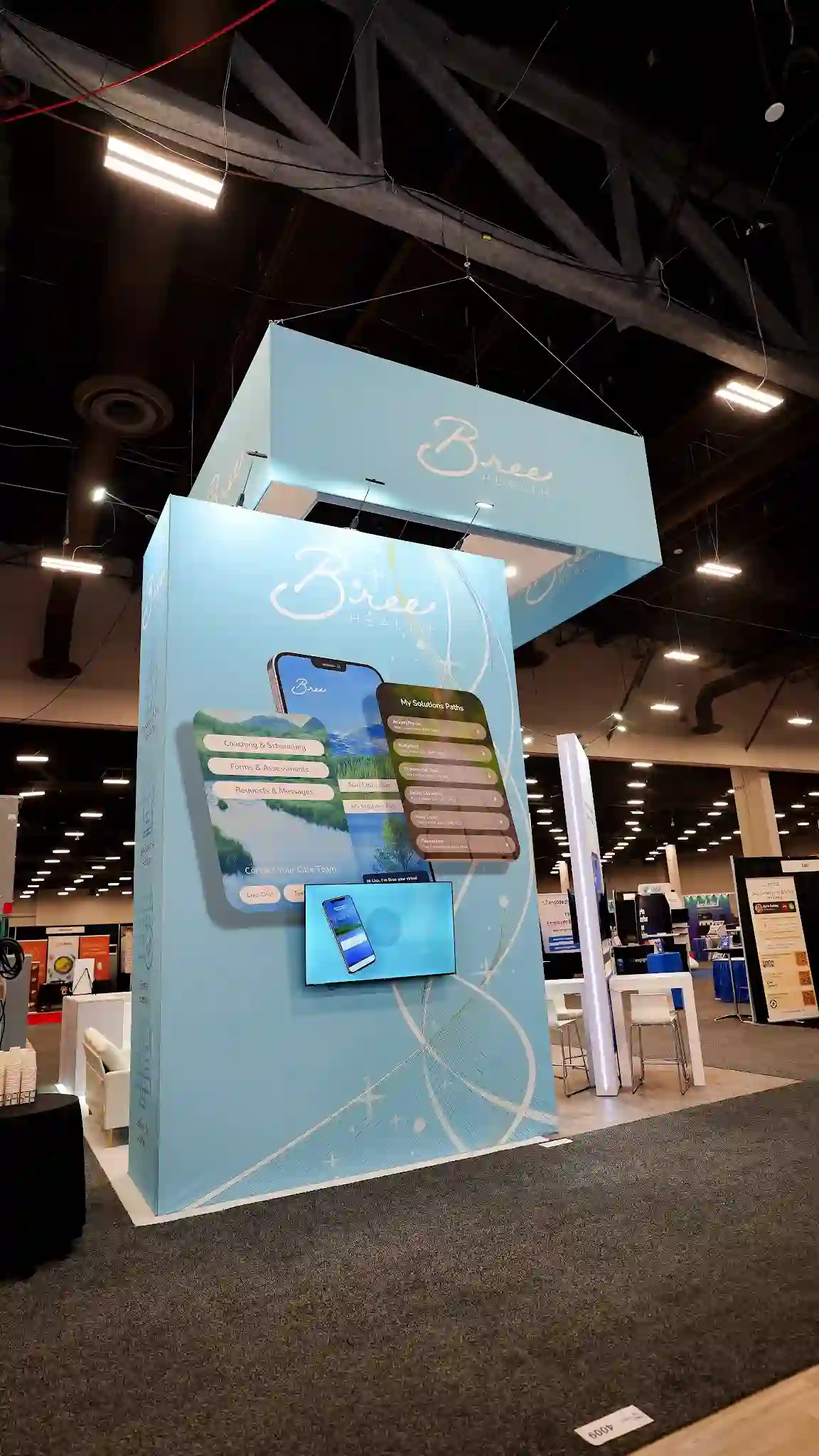 Vertical ground-level upward view of the Bree Health booth at HR Tech 2025 showing the tall central tower structure wrapped in light blue brand graphic with flowing abstract white ribbon patterns, Bree Health cursive logo near the top, large oversized phone graphic on tower front face displaying My Solutions Paths Coaching and Scheduling Forms and Appointments Payments and Messages app interface, wall-mounted flat-screen TV below showing phone with Bree app, large overhead box-shaped hanging sign visible above in sky blue with Bree cursive logo in white suspended by cables, dark convention center ceiling with exposed ductwork and fluorescent lighting creating dramatic contrast with bright blue booth, bar-height white tables and chairs partially visible within booth