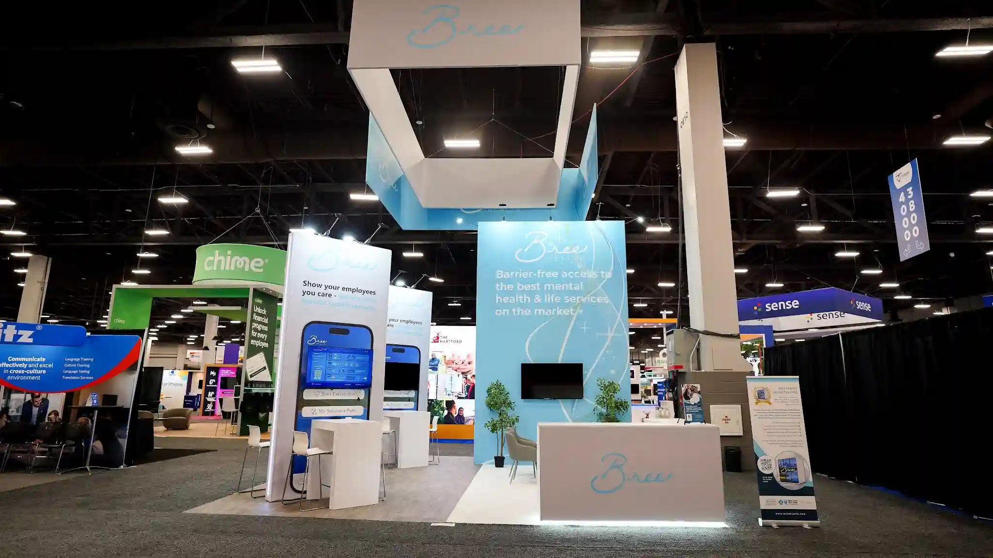 Wide-angle establishing shot of the complete Bree Health 20x30 ft island exhibit at HR Tech Conference 2025 Booth 7905 at Mandalay Bay Las Vegas showing the full booth footprint with dramatic overhead hanging sign structure in white and sky blue featuring the cursive Bree wordmark suspended from ceiling by steel cables, tall branded backwall on the right wrapped in light sky blue graphic with flowing white abstract ribbon line patterns displaying the Bree Health logo and tagline Barrier-free access to the best mental health and life services on the market, wall-mounted flat-screen TV flanked by potted green trees, gray upholstered accent chairs and white low coffee table in the lounge area, tall white interactive kiosk towers on the left displaying oversized smartphone graphics showing the Bree Health app interface with Show your employees you care messaging, white reception counter at front with LED accent strip lighting and cursive Bree logo in teal, warm wood-look vinyl flooring throughout the booth, bar-height white table and stool on left side, neighboring Chime and Sense booths visible in background, Mandalay Bay convention center dark ceiling with exposed structural beams overhead