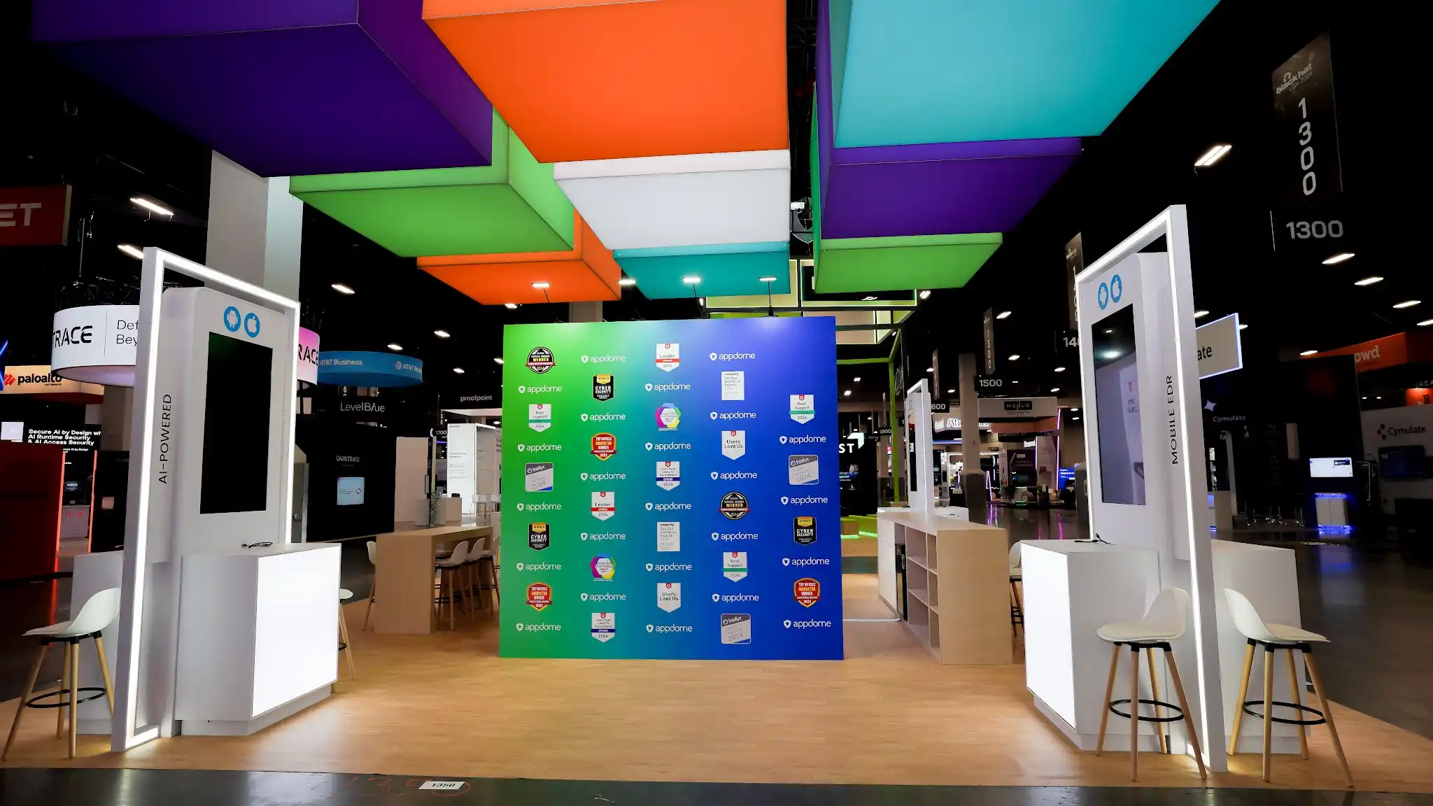 Direct frontal view of Appdome island exhibit at Black Hat USA 2024 showing awards and recognition wall with green and blue color-blocked sections displaying certification badges partner logos and industry awards in a grid pattern, overhead colorful cube sculpture visible above, white MOBILE LINK kiosk stations creating a gateway entrance, bar-height seating areas with natural wood stools, and open inviting layout contrasting with dark cybersecurity booths surrounding the exhibit