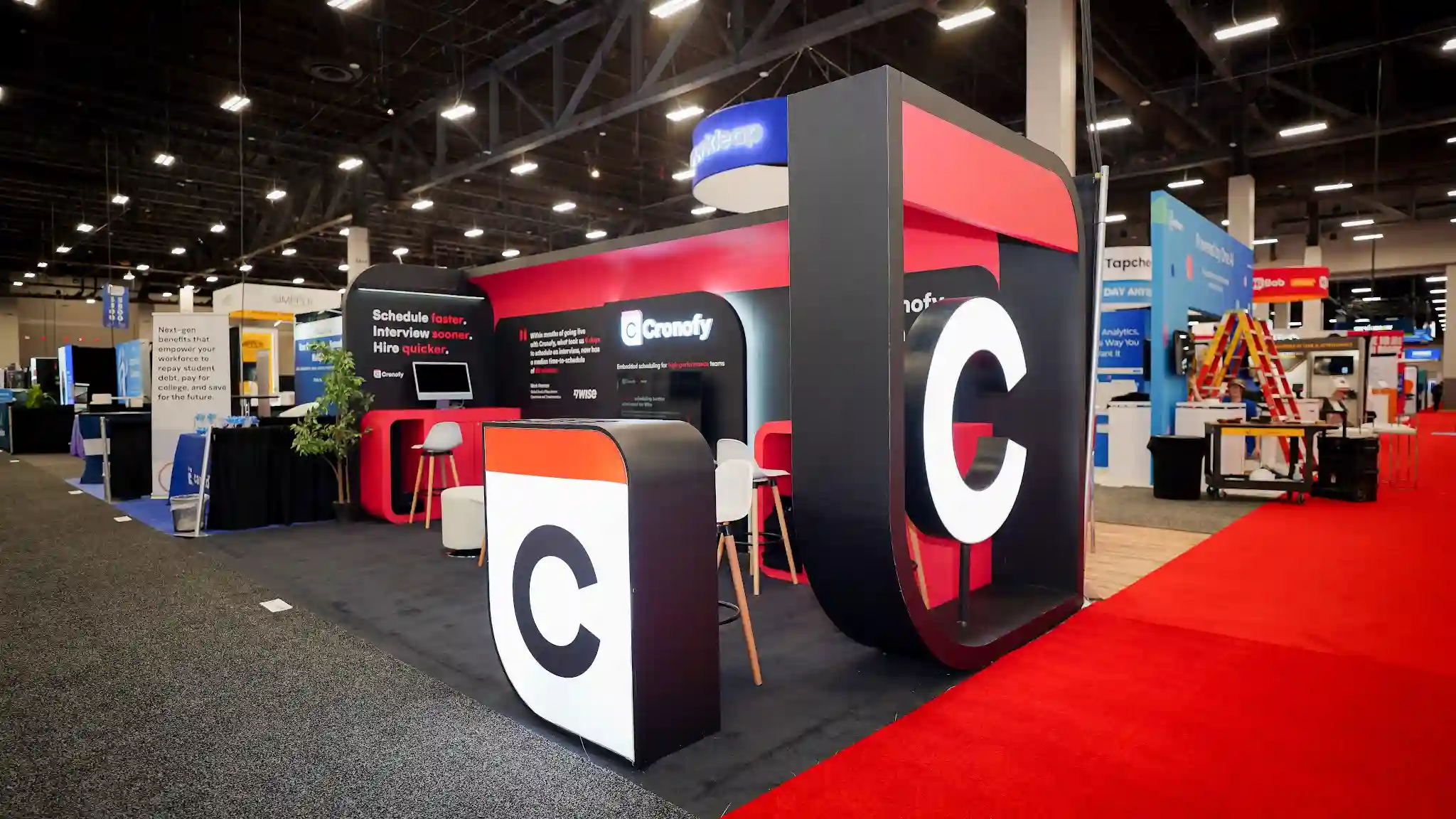 Front corner view of the Cronofy 10x20 ft island booth at HR Tech 2025 taken from the right-front corner providing a strong perspective view down the length of the booth, oversized 3D Cronofy C app icon prop prominently placed in foreground center with white face black letter and orange-red top stripe on dark black base, second taller dark vertical pillar styled as part of Cronofy icon system visible to far right, Schedule faster Interview sooner Hire quicker on left arched pod, Cronofy logo at center, two giant oversized C app icon props creating visual bookend entrance effect, arched rounded tops of flanking pod structures prominent, red carpet aisle on right adding dramatic contrast to dark booth flooring, decorative indoor tree in left pod, neighboring Tapcheck and HiBob booths visible in background