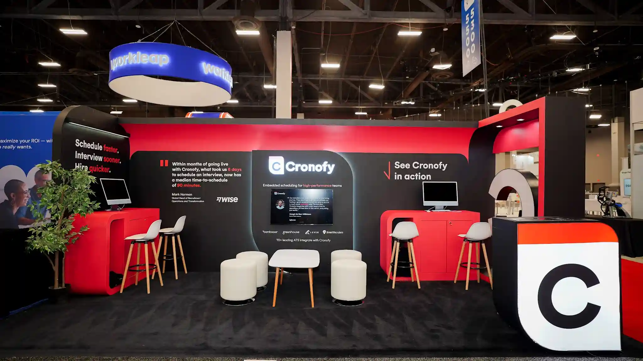 Straight-on front view of the complete Cronofy 10x20 ft custom island exhibit at HR Tech Conference 2025 Booth 3217 at Mandalay Bay Convention Center Las Vegas showing the full booth structure with bold red black and white color palette, central back wall featuring Cronofy logo and Embedded scheduling for high-performance teams tagline, Schedule faster Interview sooner Hire quicker headline on left panel with Wise customer testimonial from Mark Harman, 70 plus ATS integration partner logos including BambooHR Greenhouse Lever SmartRecruiters, See Cronofy in action call-to-action on right panel, two rounded arch portal entryway structures with curved frames on left and right flanks, oversized 3D C app icon prop on far right, central lounge seating with white round ottomans and coffee table, red lacquered countertops with iMac demo stations, bar stool seating, decorative potted tree inside left pod, dark charcoal carpet flooring, neighboring Workleap booth with blue illuminated sign visible