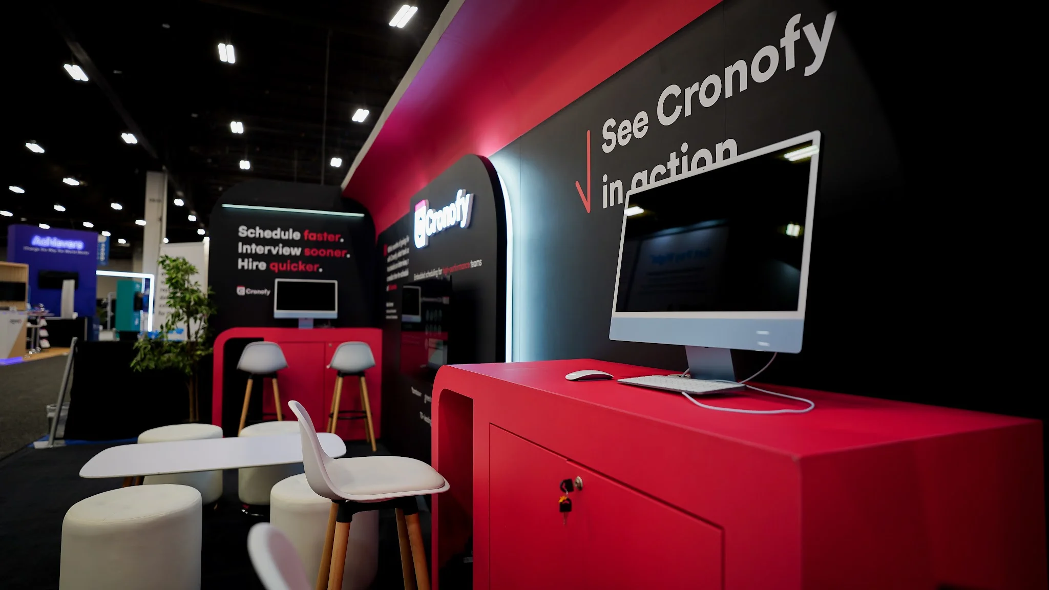 Close-up interior view of the Cronofy booth at HR Tech 2025 focused on the right-side demo station showing vivid red lacquered presentation counter with lockable cabinet below as focal point, Apple iMac white silver computer set up on red counter with keyboard and mouse for live product demos, See Cronofy in action in large white lettering on right back wall panel with bold red downward-pointing arrow call-to-action, Cronofy logo with icon on central back wall, Schedule faster Interview sooner Hire quicker on left back panel, Embedded scheduling for high-performance teams sub-tagline visible, LED-lit curved arch frame casting soft glow adding high-tech premium feel, dark ceiling interior with bright red and white elements, decorative potted plant faintly visible in background