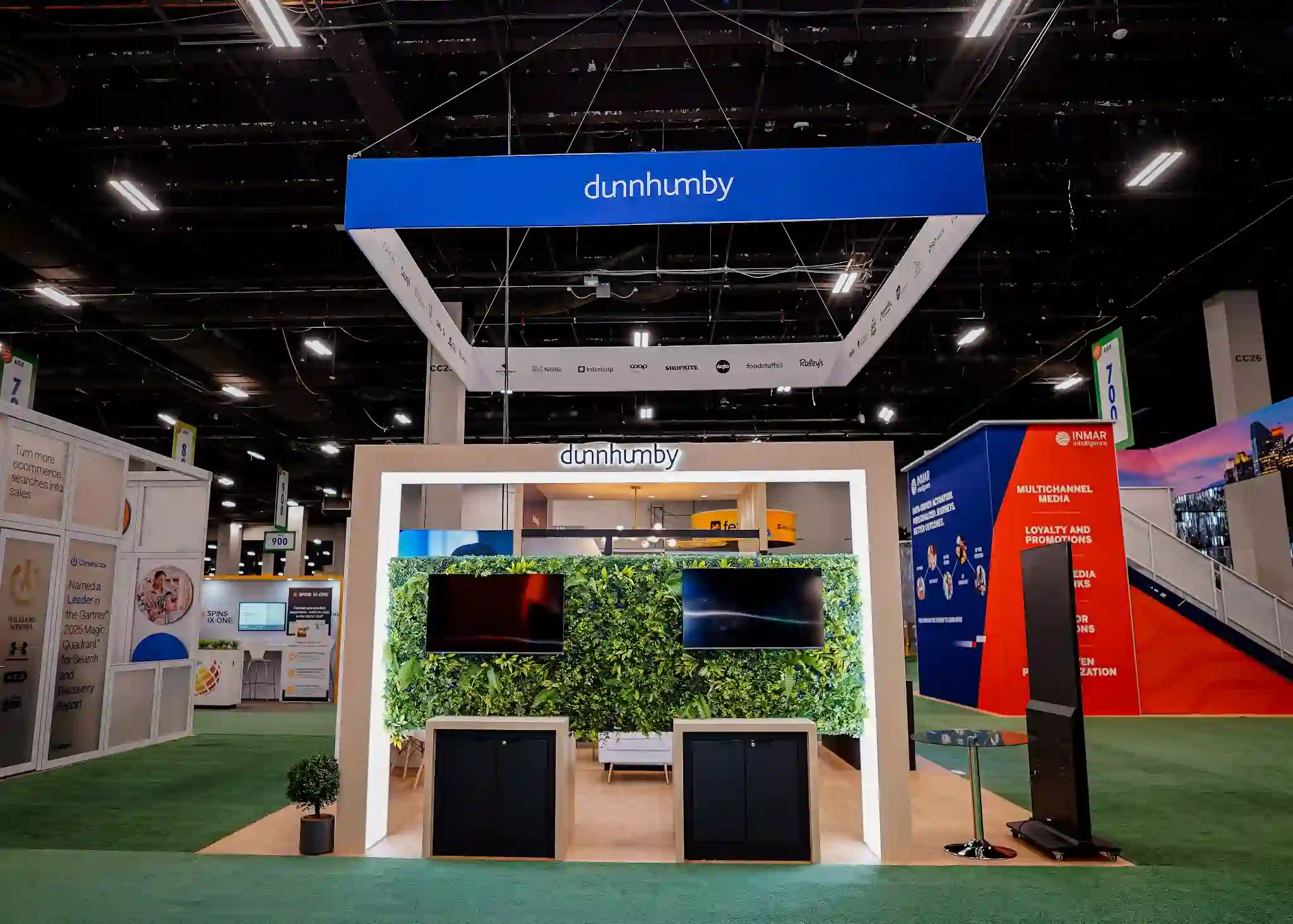 Full-height living green wall with dense foliage, backlit dunnhumby logo above center, two flat-screen monitors embedded in greenery showing high-contrast content, white architectural frame, and black minimalist equipment housings below
