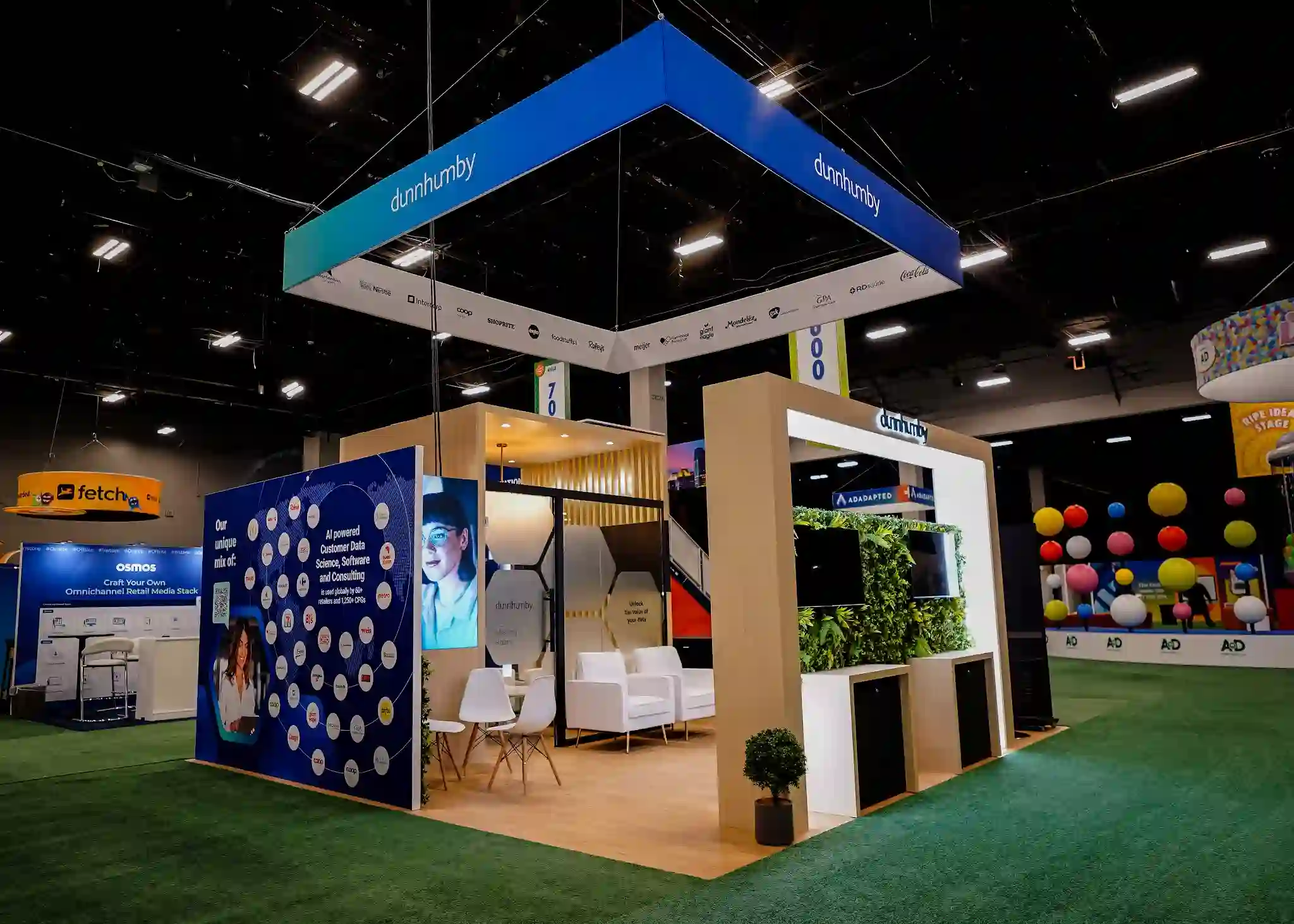 dunnhumby booth side view showing blue graphic wall with partner brand logos web including L'Oreal Coca-Cola and Nestle, Our Unique Mix Of header text, white mid-century modern lounge chairs, round meeting table, and vertical wood slat screens with greenery