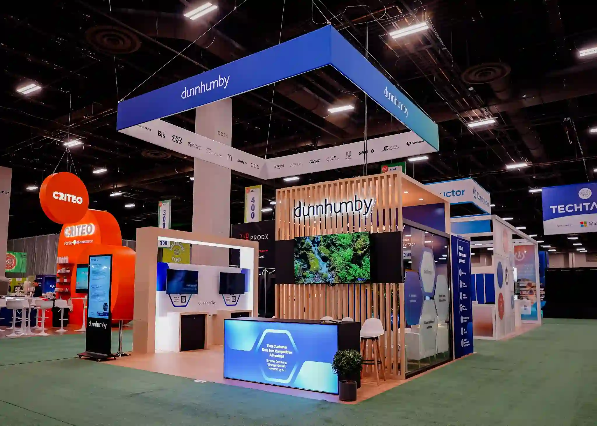 dunnhumby reception counter with blue backlit front panel reading Turn Customer Data into Competitive Advantage, monitor on wood slat wall displaying nature waterfall scene, backlit dunnhumby logo with 3D effect on vertical wood slats, and blue LED accent lighting