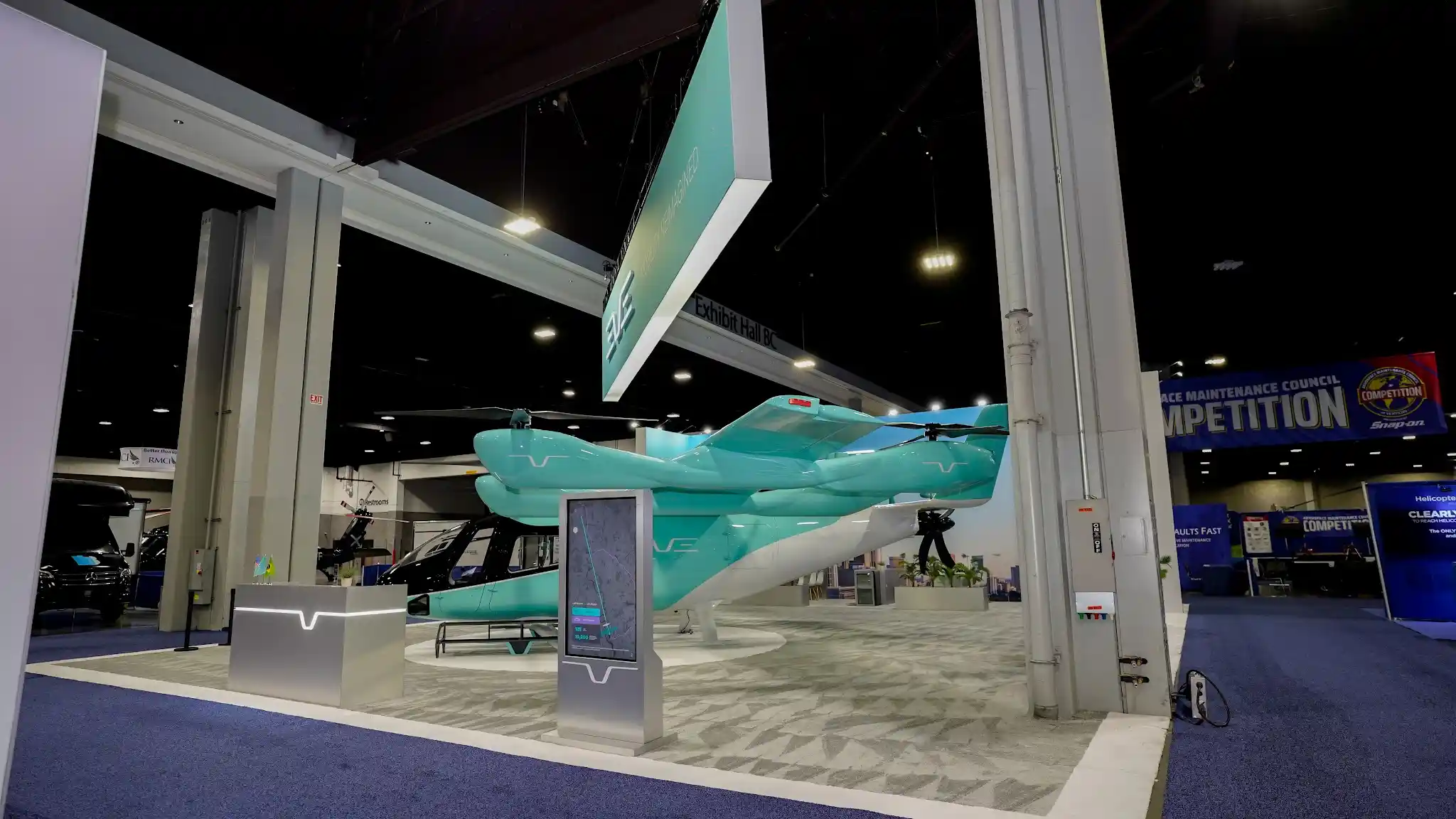 Side angle view of turquoise EVE eVTOL aircraft with white and silver belly, eight-rotor configuration, black canopy, white architectural pillar with integrated EVE logo lighting, and gray display kiosk with screen at VertiCon 2026