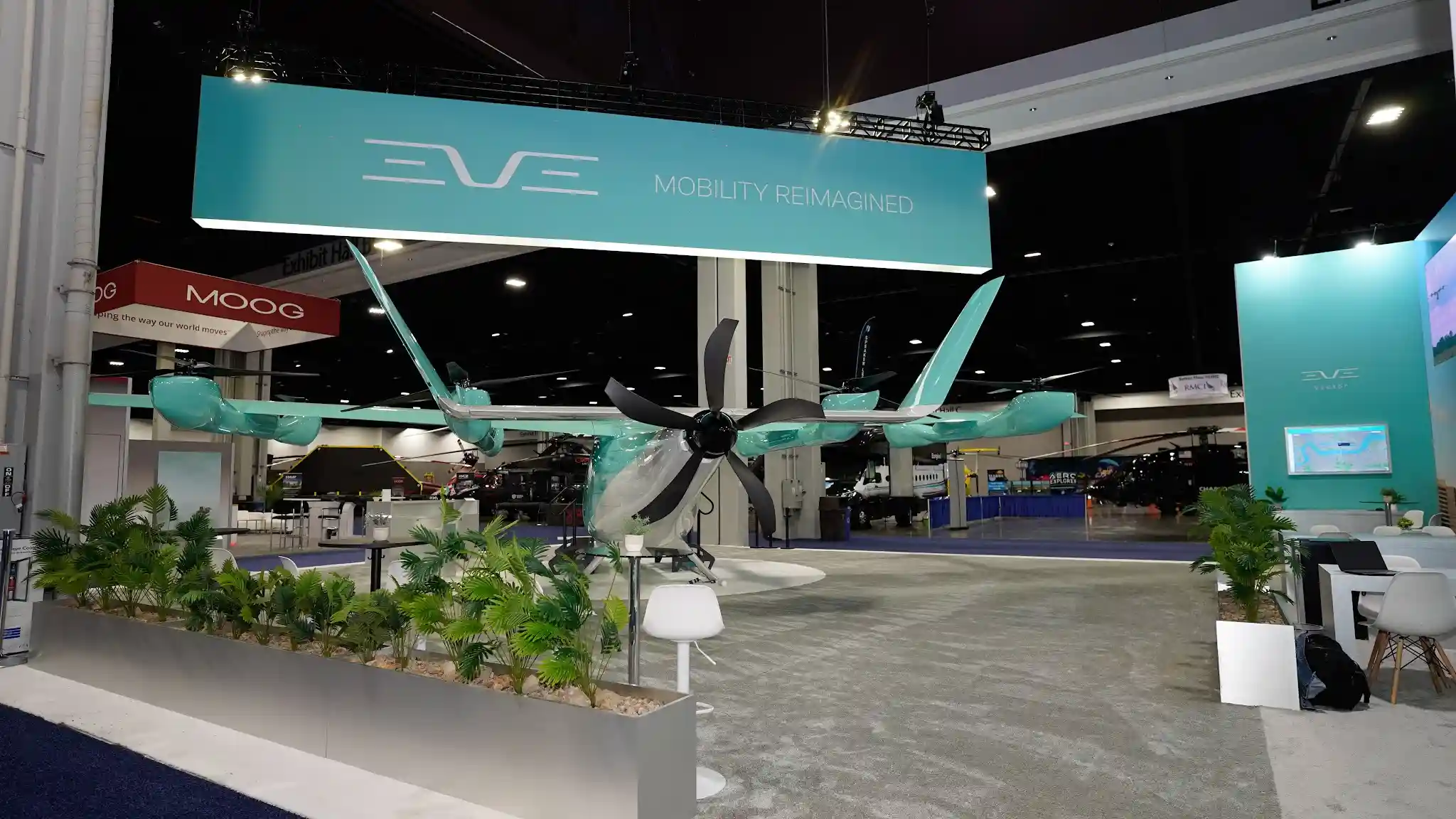 Expansive wide-angle view of full booth layout with eVTOL aircraft, extensive greenery in white planters, turquoise display panel with screen, low white perimeter barriers, and open floor plan
