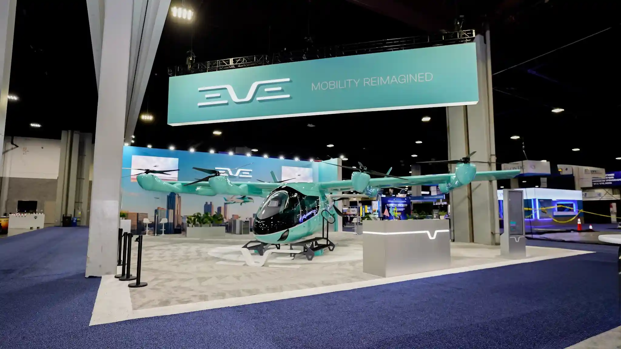 Full-scale EVE eVTOL aircraft mock-up in turquoise with eight rotors and black canopy, mounted on white display platform, EVE Mobility Reimagined overhead hanging sign, black stanchion barriers, urban skyline backdrop graphics at VertiCon 2026