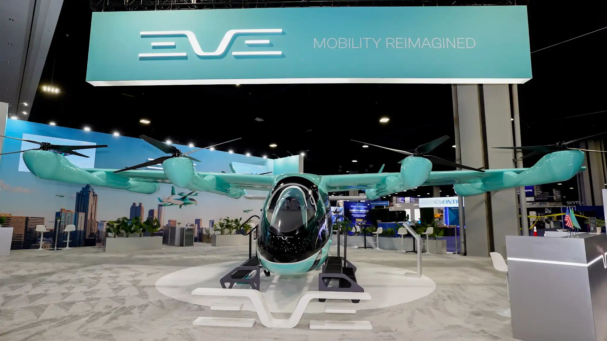 Front view of EVE Air Mobility 45x55 custom island exhibit at VertiCon 2026 with full-scale turquoise eVTOL aircraft on white sculptured display platform, EVE Mobility Reimagined overhead hanging sign, urban skyline graphics, white modern furniture, and tropical plants at Georgia World Congress Center in Atlanta