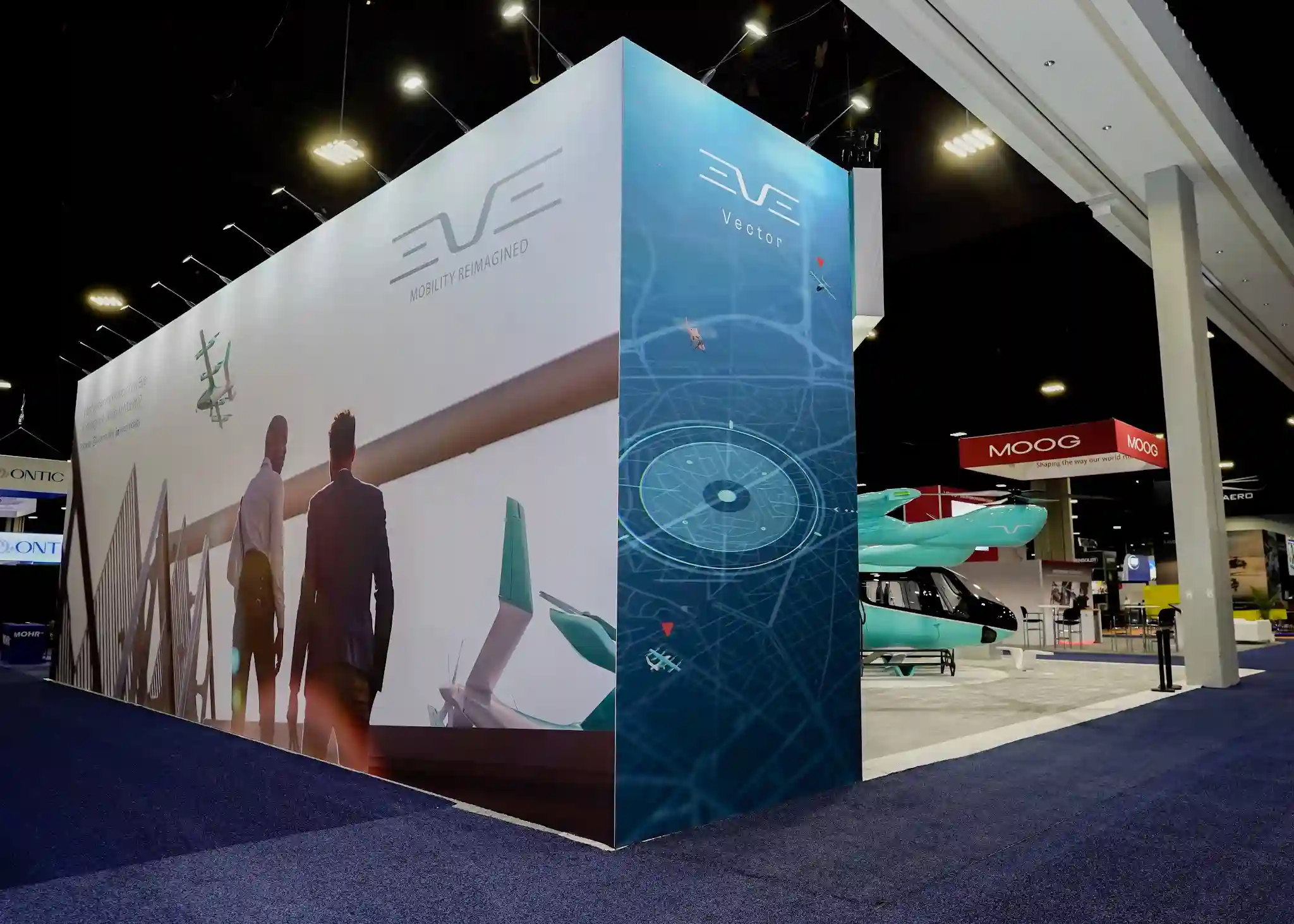 EVE Vector corner booth with dramatic three-dimensional turquoise pillar featuring digital radar and navigation interface graphics, EVE Mobility Reimagined wall text, lifestyle photography of silhouetted figures at sunset viewing aircraft, and physical turquoise eVTOL model with red accents visible in interior