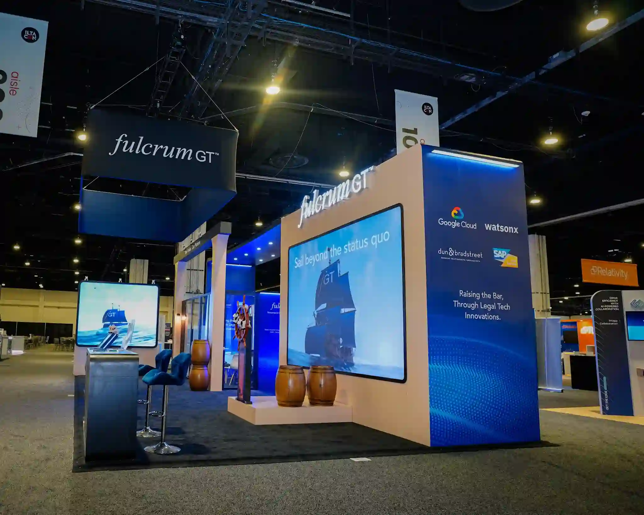 Massive LED screen displaying sailboat and Sail beyond the status quo messaging, blue partner wall listing Google Cloud Watsonx Dun and Bradstreet and SAP with Raising the Bar Through Legal Tech Innovations headline, black rectangular hanging sign with white fulcrum GT lettering, demo station with blue bar stools, and dark charcoal carpeting