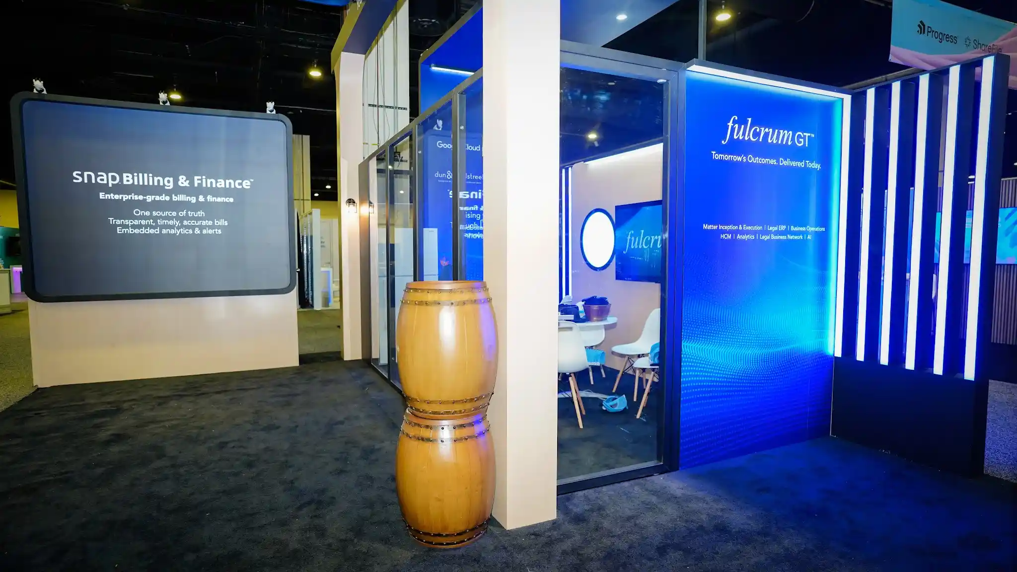 Semi-enclosed meeting area with glass walls, large screen displaying snap Billing and Finance Enterprise-grade billing and finance with value propositions, backlit deep blue wall with fulcrum GT logo and Tomorrows Outcomes Delivered Today tagline, blue glowing dot pattern, wooden barrel nautical decor