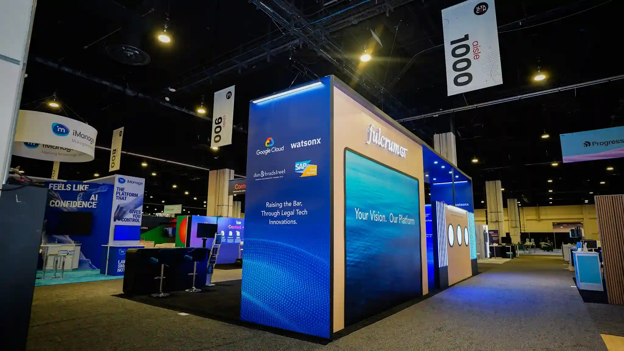 Fulcrum GT island booth viewed from Aisle 1000 showing vertical slatted wall with white beams and blue backlighting, tower screen displaying Your Vision Our Platform, blue LED ceiling panels, and dark charcoal carpeting with iManage and Progress booths visible in background
