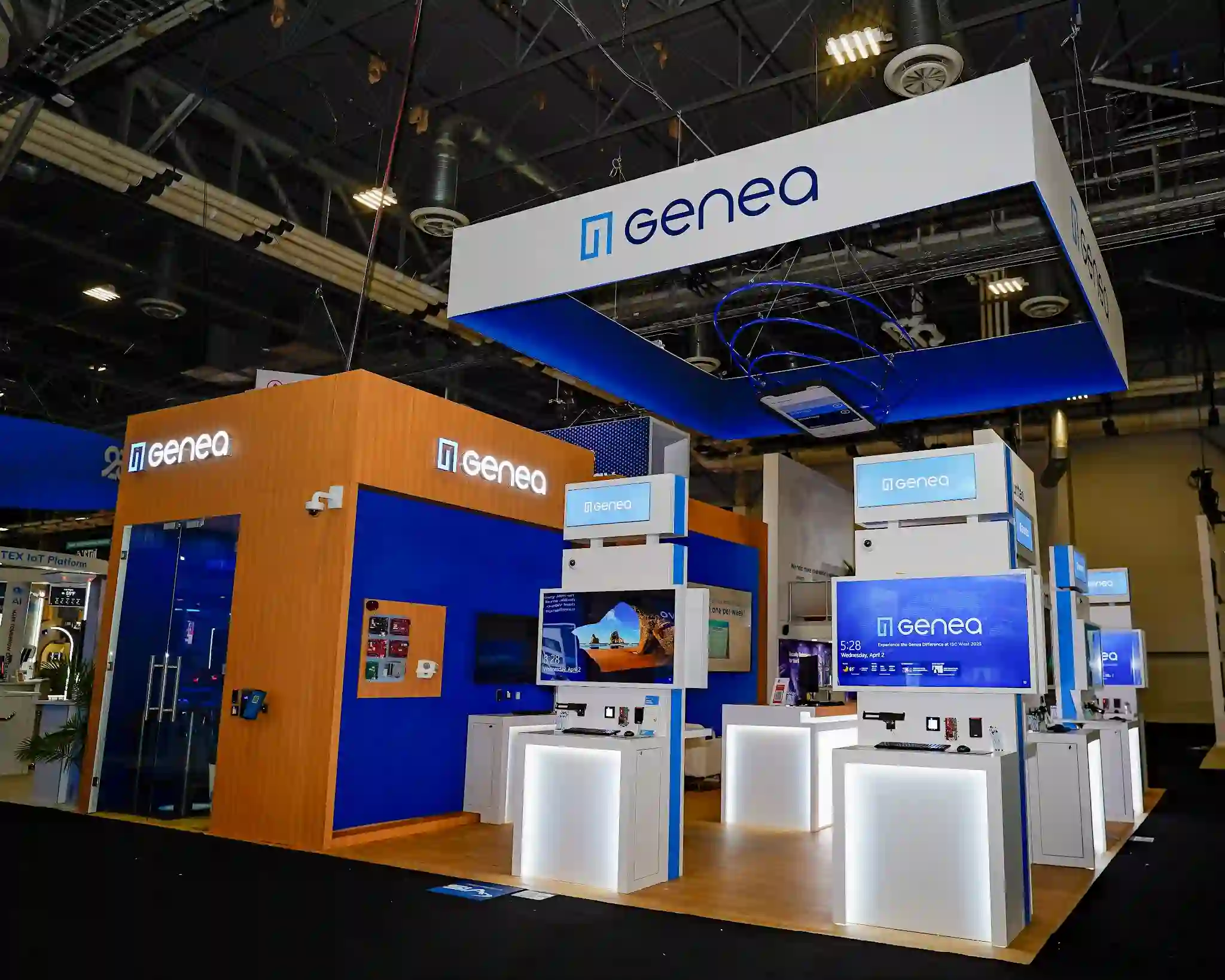 Transition zone between blue demo kiosks and warm wood meeting area, Genea logo on kiosk tops and orange meeting structure, Hardware Integration wall visible on blue surface