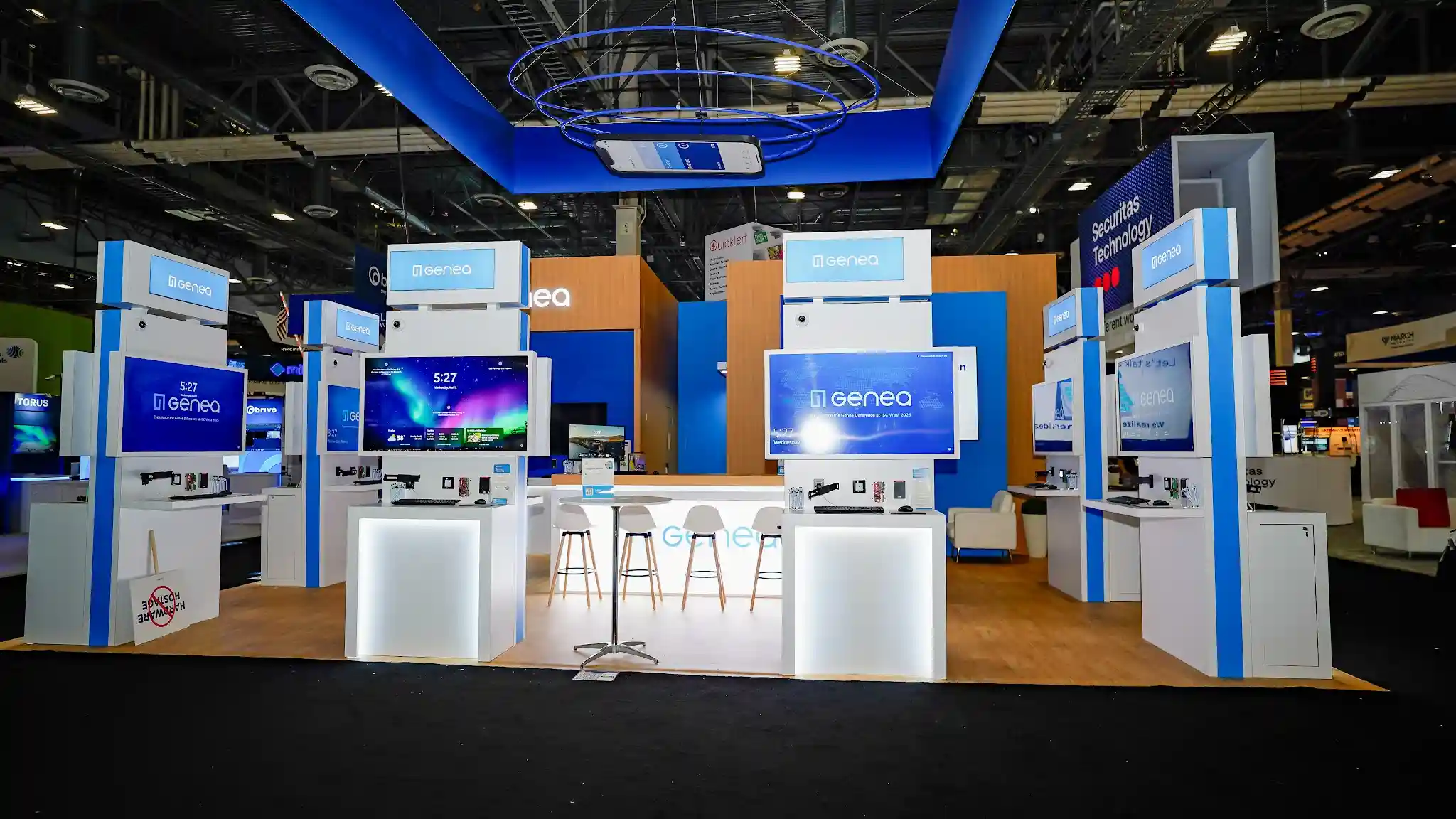Central networking area with suspended mobile-phone-shaped display, white circular cocktail table with bar stools, Securitas Technology pylon in background, and multiple kiosks showing Genea mobile app
