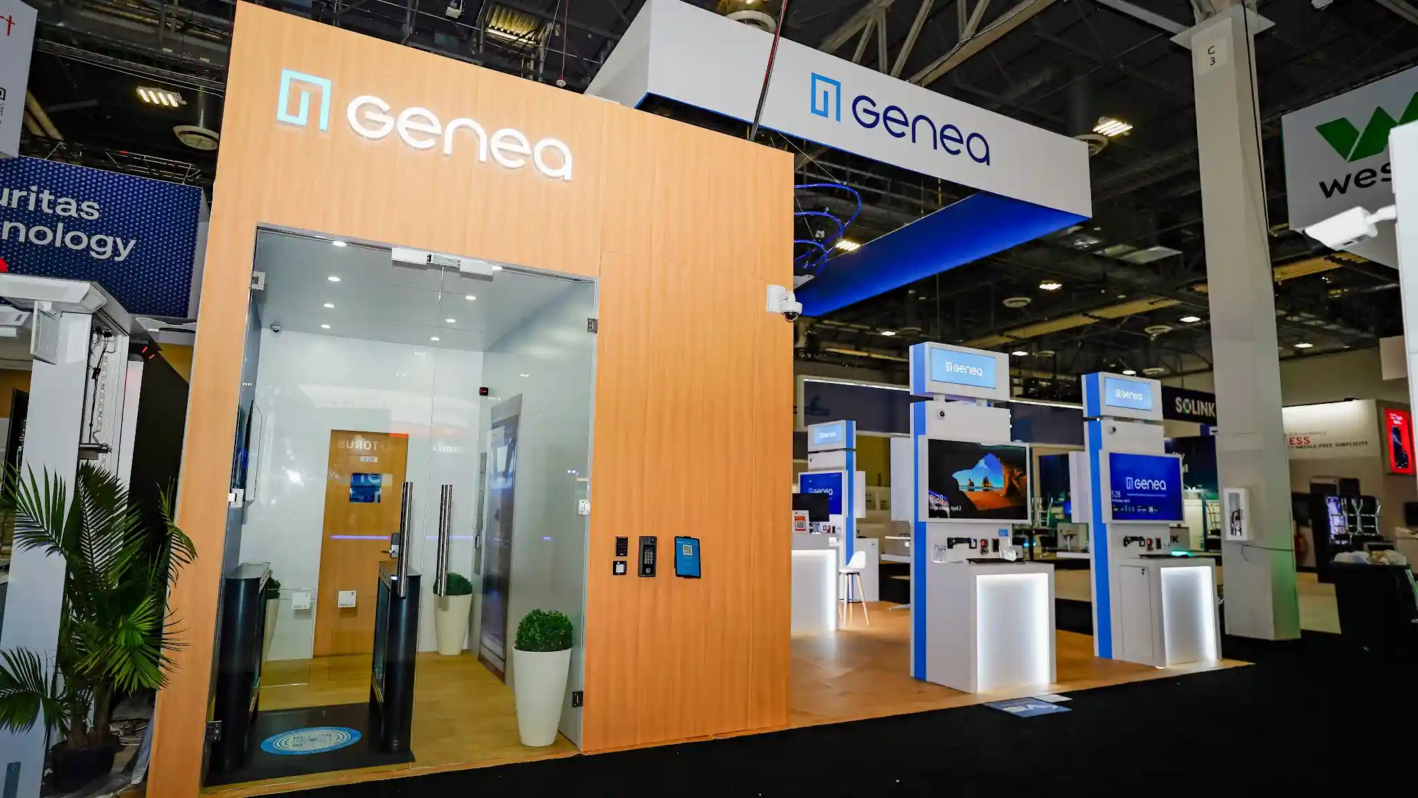Genea Security Experience room featuring light wood exterior with backlit Genea logo, large glass entry door, optical turnstile inside, security camera mounted on top corner, access control readers at chest height for interactive testing, recessed puck lighting, and clean white metal trim
