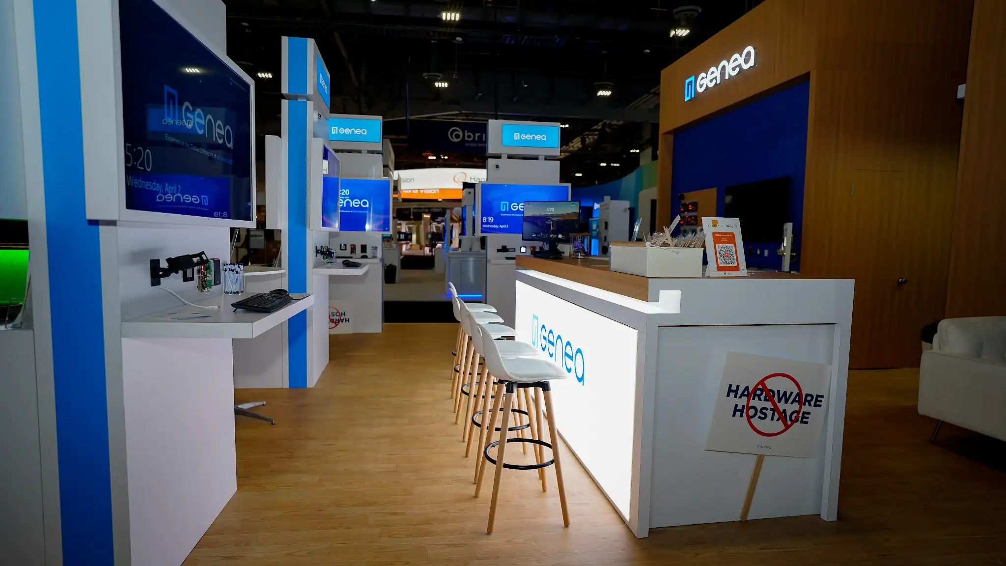 L-shaped reception counter with bright white backlit Genea logo, natural wood-grain top, five white bar stools with wood legs, No Hardware Hostage campaign sign with red prohibition symbol, and blue-themed demo screens in background