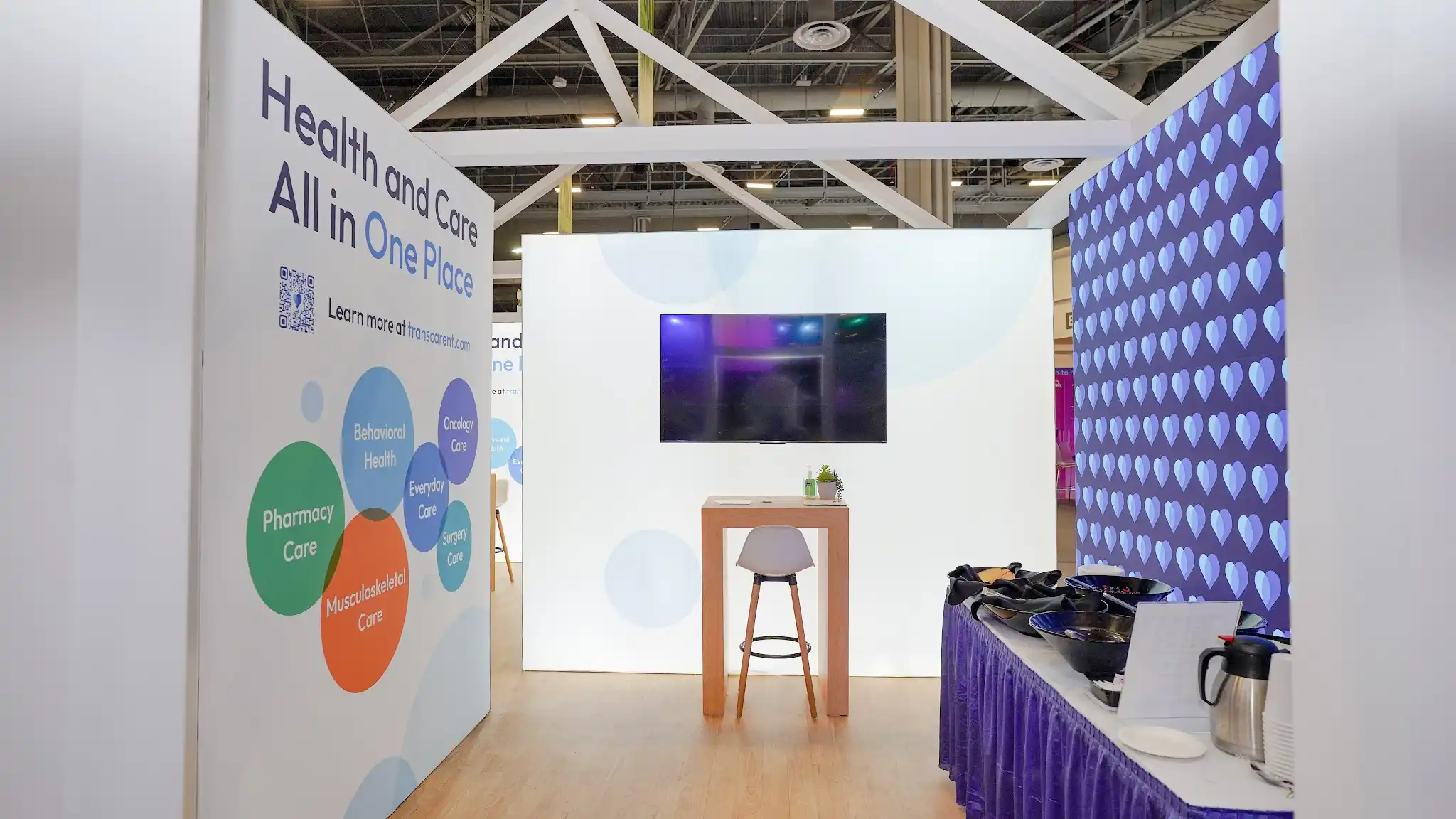 Interior perspective of the Transcarent house booth at HLTH 2023 looking toward back and sides showing deep navy blue heart-patterned interior accent wall on right with repeating lighter blue 3D heart shapes functioning as branded wallpaper, white backlit rear panel wall with TV monitor mounted at center, wooden counter-height table with modern white chair on wooden barstool base and small green succulent plant, purple-skirted hospitality refreshment table with black thermos coffee dispensers on far right, warm honey-blonde wood-look laminate plank flooring, white aluminum structural frame columns as architectural elements, front interior panel angled showing Health and Care All in One Place messaging with QR code and colorful bubble service diagram partially visible