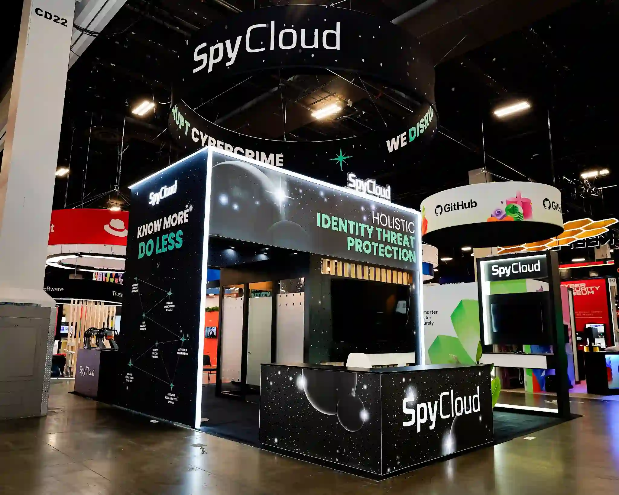 Front corner angle view of SpyCloud 20x20 island exhibit at Black Hat USA 2025 showing rectangular two-story structure with cosmic starfield graphic panels, Holistic Identity Threat Protection headline and Know More Do Less side panel in teal and white, large circular overhead hanging sign with SpyCloud logo and We Disrupt Cybercrime ring text, white LED edge lighting creating glowing halo effect on all structural edges, reception counter with SpyCloud branding at front, white bar stools inside, green tropical plant in white pot, vertical natural wood slat accents visible through interior opening, and neighboring GitHub and Red Hat booths in background