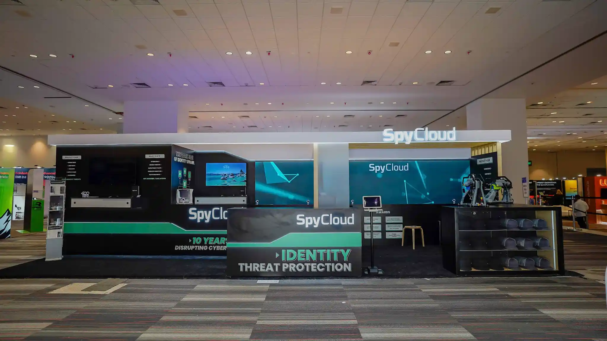 Full-width head-on frontal view of the SpyCloud 10x30 ft custom booth at RSA Conference 2026 Booth S-3338 in Moscone Center South Hall, showing the complete dual-zone layout divided into a left demo and content zone and a right engagement and merchandise zone connected by a center walkthrough aisle, the illuminated white channel-letter SpyCloud logo glowing on the white canopy header as a wayfinding beacon visible from across the show floor, matte black structural panels creating the dominant dark-tech backdrop, teal-lit LED backwall with geometric network visualization motifs as the interior centerpiece, two collaborative robotic arms positioned in the right engagement zone serving as an interactive technology attraction drawing attendees, glass merchandise display case with stacked SpyCloud branded hats elevated to retail-quality presentation, awards display panel framed with industry recognition certificates, dark charcoal custom flooring cleanly demarcating the SpyCloud footprint from the surrounding convention carpet, multiple large demo monitors and tablets running live platform demonstrations, RSA Conference 2026 Moscone Center South Hall environment visible in background