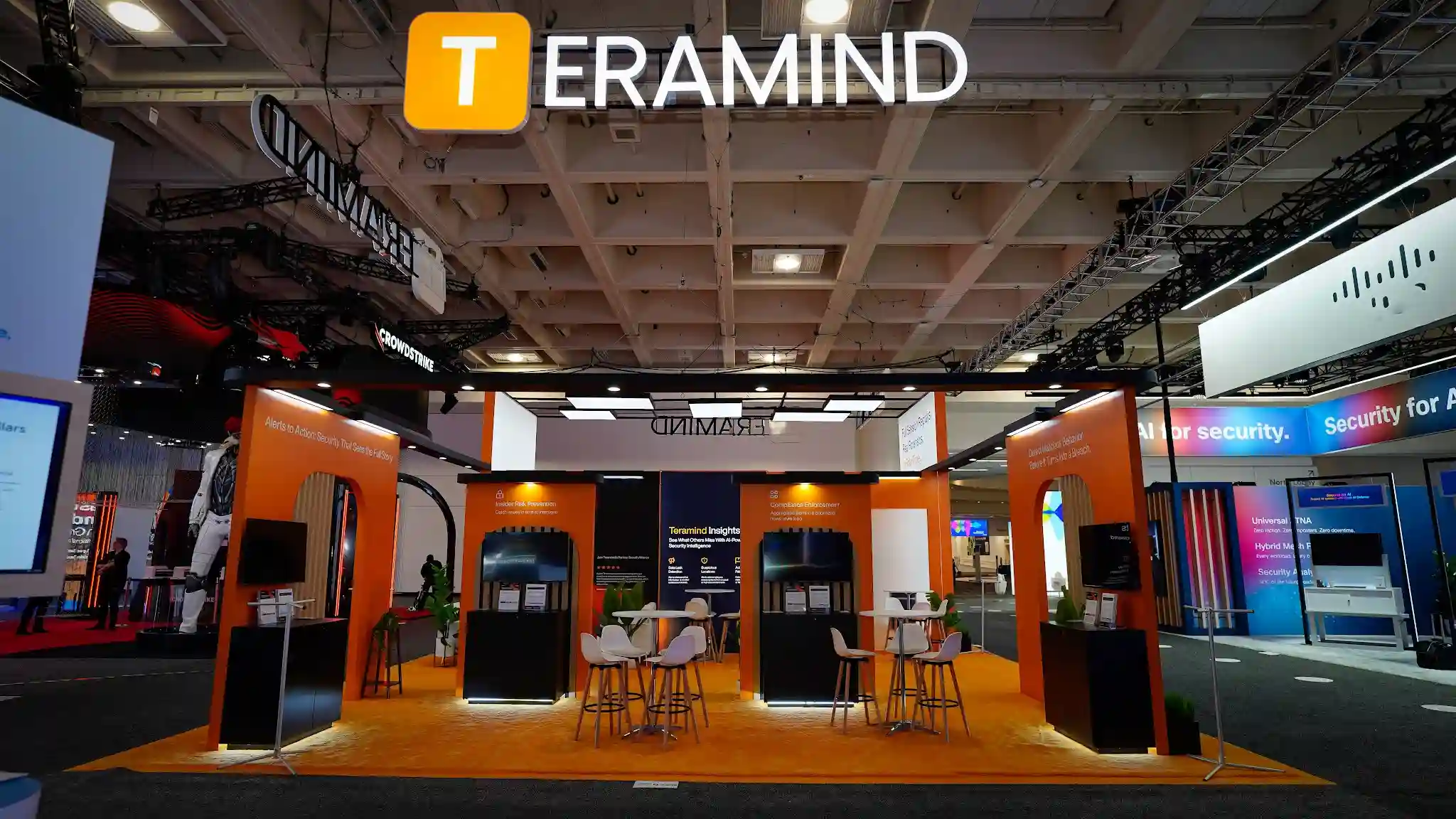 Interior kiosk gallery view of the Teramind booth at RSA Conference 2025 showing multiple product demo monitors displaying Teramind platform screens and cybersecurity analytics dashboards, product messaging panels on the walls reading Investigate Threats 65 Percent Faster and Every Action Every User Full Attribution, burnt-orange accent lighting and panels, 5-star customer review graphic visible, amber carpet flooring, LED ceiling panel tiles