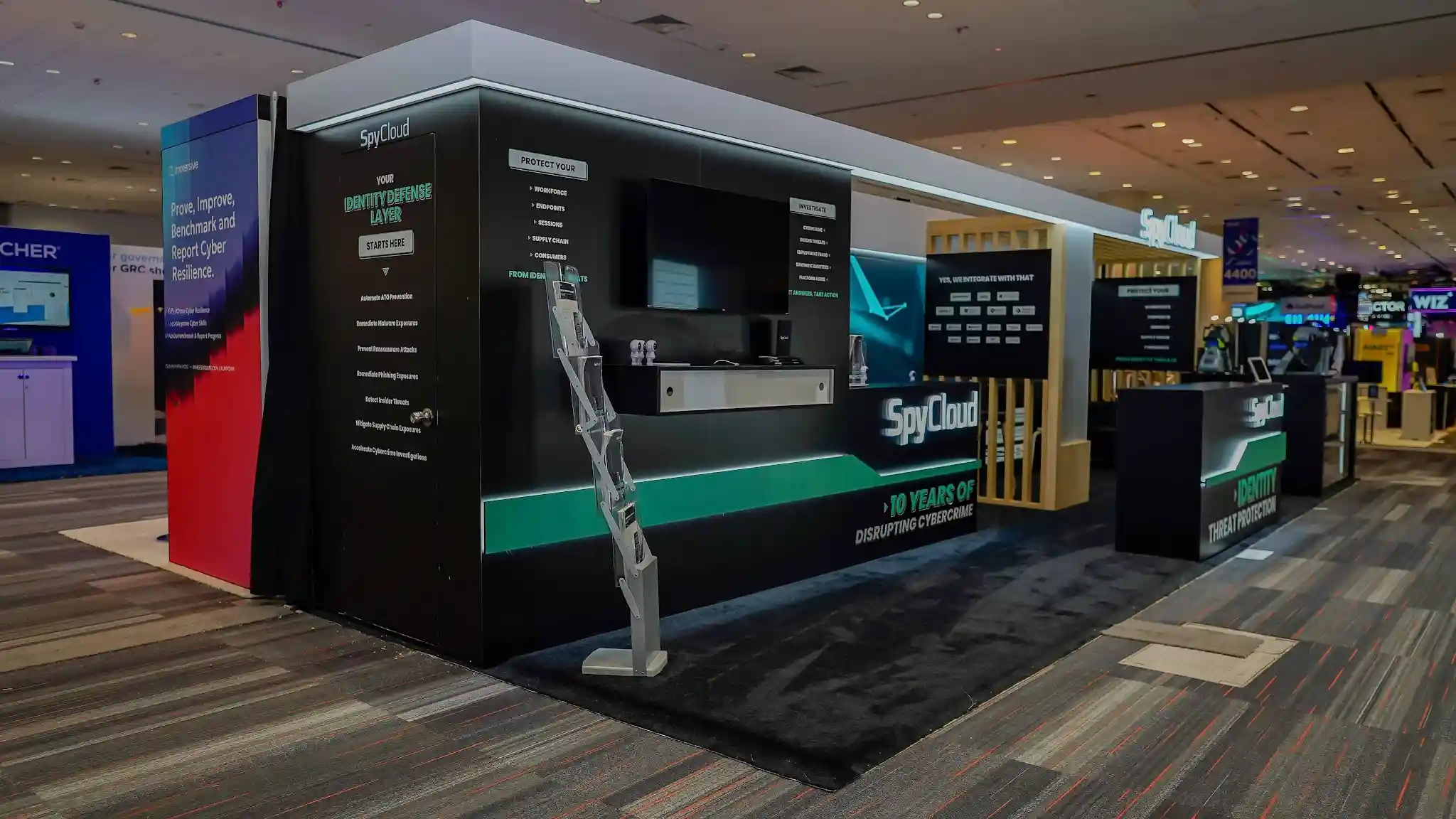 Left-front three-quarter angle view of the SpyCloud 10x30 ft custom booth at RSA Conference 2026 showing the main demo and content wall with Your Identity Defense Layer Starts Here headline messaging in bold white typography on matte black panels, Protect Your Workforce Endpoints Sessions Supply Chain and Consumers product capability framework displayed prominently, 10 Years of Disrupting Cybercrime milestone messaging on the counter fascia with teal accent stripe, natural blonde vertical wood slat architectural column serving as warm organic counterpoint to the dark tech palette, multi-tier literature rack for brochure and leave-behind distribution, large monitor running live SpyCloud platform demonstration, teal accent details and SpyCloud branded arrow motifs providing visual wayfinding across the booth structure