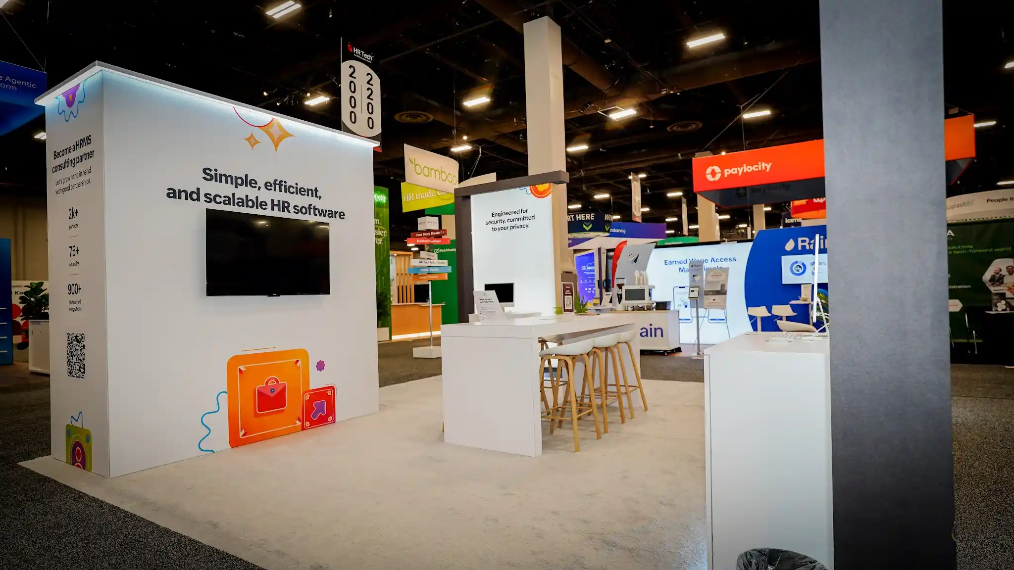 Interior view of the Zoho booth showing bar seating area with orange stools, partner stats panel, central product display with flat-screen TV, and wayfinding signage