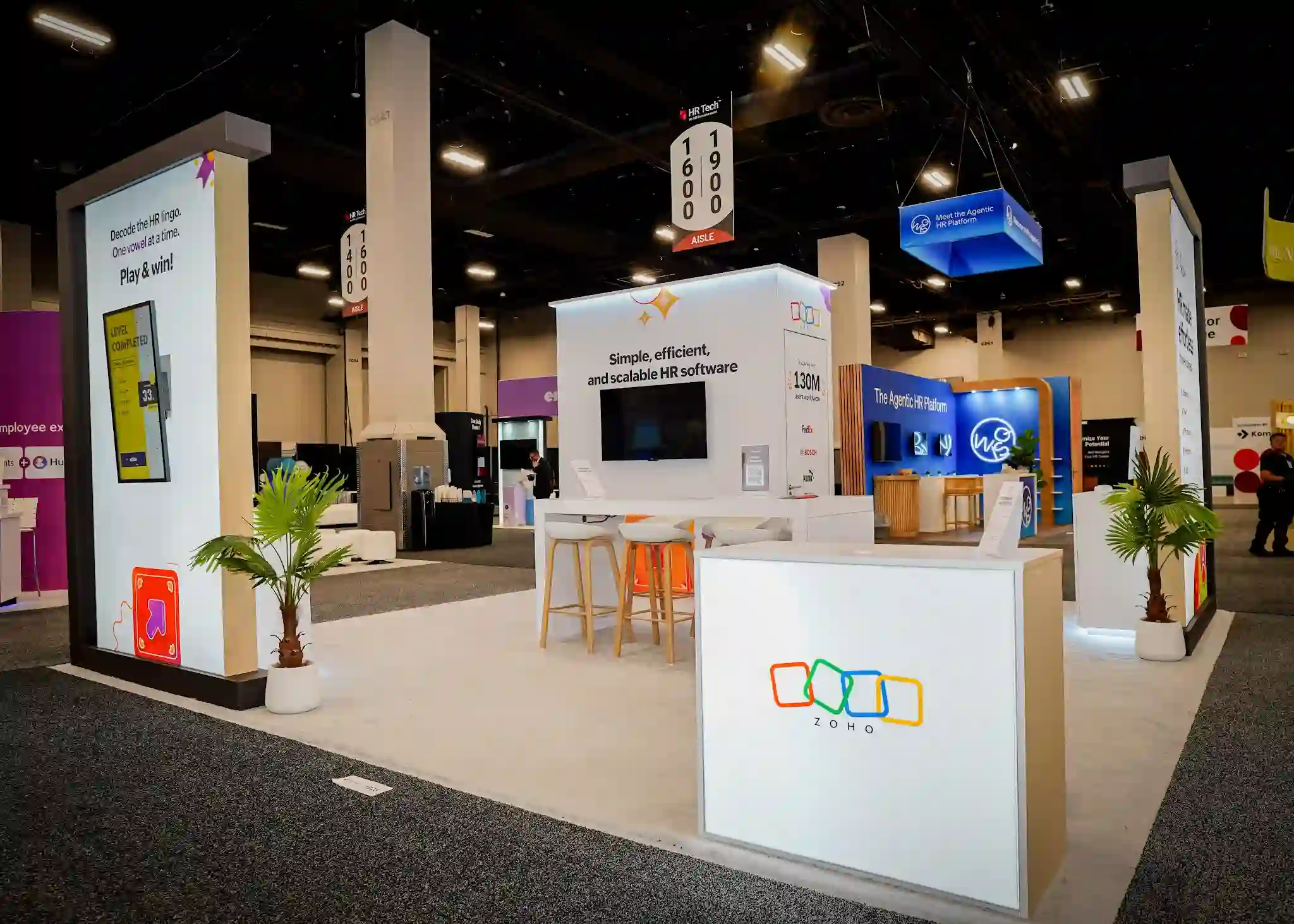Wide-angle frontal overview of the full Zoho island booth at HR Tech 2025 showing all four dark-charcoal-framed rectangular gateway towers framing brilliant white backlit lightbox panels, interactive gaming display with yellow Level Completed screen, central product messaging column, Zoho People feature panel, backlit Zoho logo reception counter, orange bar stools at white tables, tropical palm plants in white planters, and bright white custom flooring