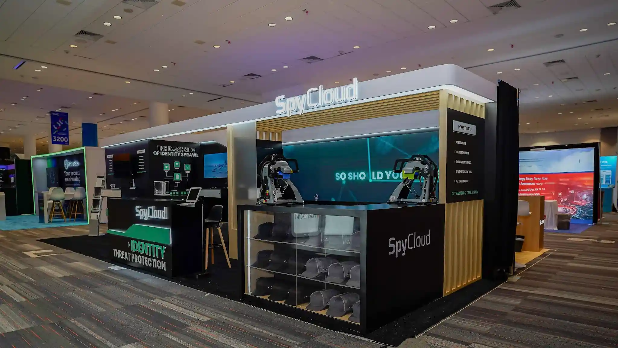 Right-front three-quarter angle view of the SpyCloud booth at RSA Conference 2026 showing the engagement zone featuring two collaborative robotic arms in the foreground serving as interactive technology demonstration and visual crowd attraction, glass merchandise display case with stacked SpyCloud branded hats presented at retail quality in the right corner, natural blonde wood vertical slat architectural column creating warm organic contrast against the matte black structural panels, teal backlit LED wall with geometric network and data visualization motifs glowing as the interior visual centerpiece with So Should You messaging in white typography, Investigate panel listing Cybercrime Insider Threats Employment Fraud Synthetic Identities and Platform Abuse product capabilities, Scandinavian-style bar height stools with natural wood legs for conversational seating, dark charcoal custom flooring, teal accent stripe and SpyCloud arrow motifs on counter fascia