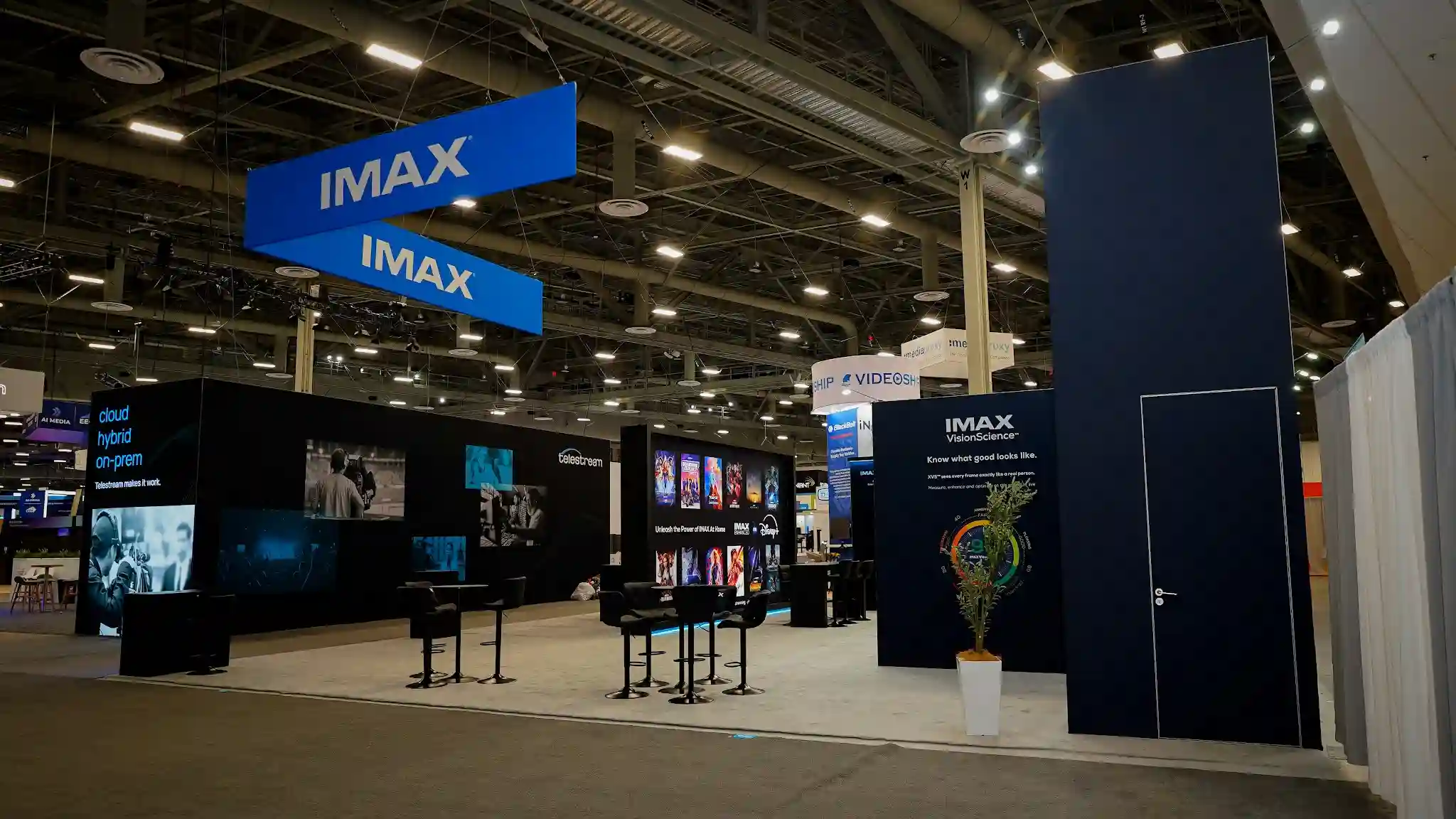 Wide front-right view of IMAX 40x50 island exhibit at NAB 2025 showing the darkest palette perspective with prominent two-tier hanging signage in deep navy and electric cyan, black-and-white photography displays, cinematic presentation screens, glass-walled rooms, and the full architectural scope of the exhibit on the Las Vegas Convention Center show floor