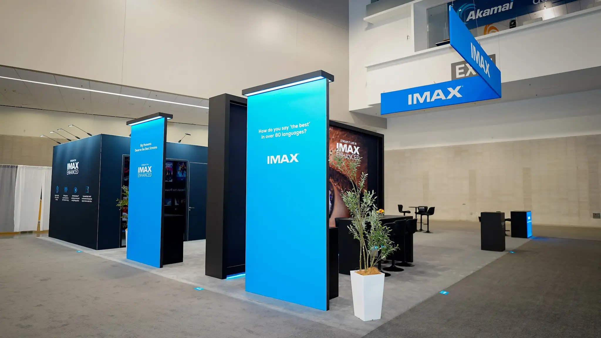 Interior corridor of IMAX exhibit showing modular cyan display panels with How do you say the best in over 80 languages messaging, dark navy walls creating immersive light-controlled environment, electric blue backlit accents, and cinematic atmosphere at NAB 2025