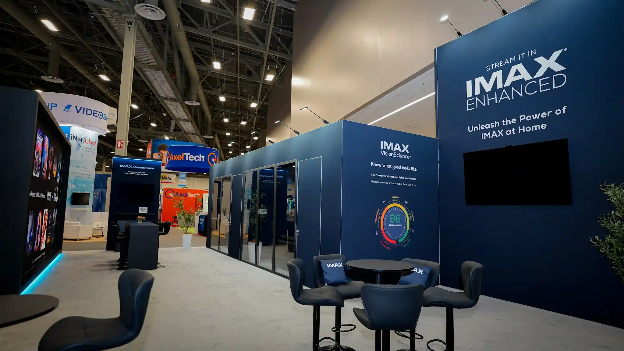 Interior of IMAX exhibit showing large display screen with cinematic content, glass-walled demonstration rooms with interior lighting, navy lounge seating with branded IMAX pillows, VisionScience wall display in background, dark cinematic walls with electric blue accent lighting, and warm ambient overhead illumination