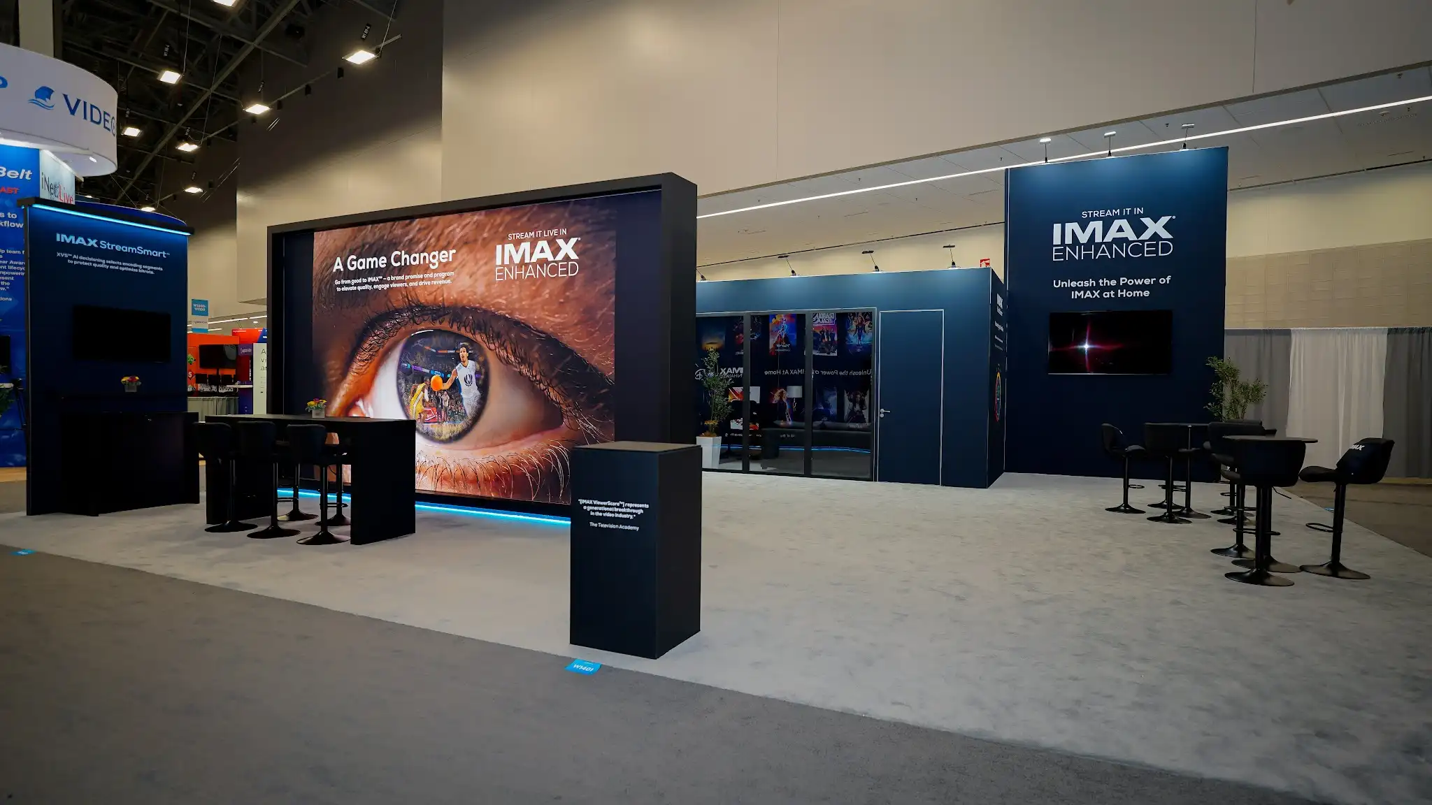 Corner view of IMAX exhibit featuring dramatic human eye imagery focal point with A Game Changer messaging, glass-walled demonstration rooms, STREAM IT LIVE IN messaging panels, movie poster displays showing IMAX Enhanced titles, and dark cinematic environment at NAB 2025