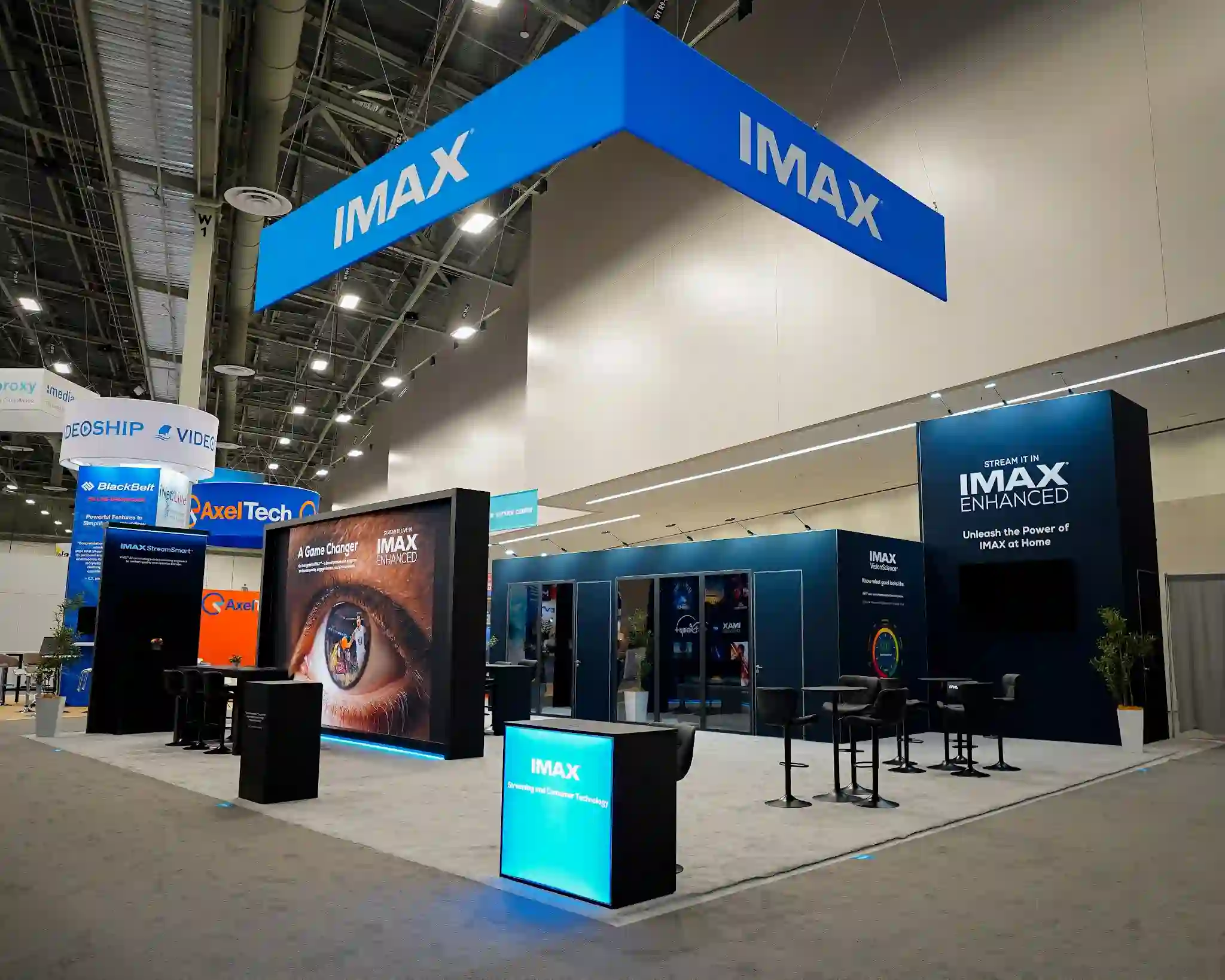 IMAX 40x50 custom island exhibit at NAB 2025 showing wide corner view with overhead IMAX two-tier hanging sign in deep navy and electric cyan, dramatic eye imagery A Game Changer display at focal corner, glass-walled meeting rooms with interior lighting, multiple engagement zones, dark cinematic walls, and blue-lit entrance portal at Booth W1401 in the Las Vegas Convention Center