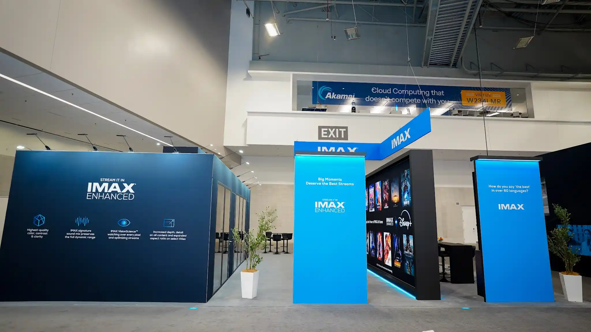 IMAX exhibit entrance view showing angular overhead IMAX sign with two-tier structure, cyan LED-lit entrance portal, feature icons for color and contrast, immersive sound, expanded aspect ratio, and enhanced detail, streaming technology messaging panels, and dark cinematic walls creating an immersive threshold experience at NAB 2025