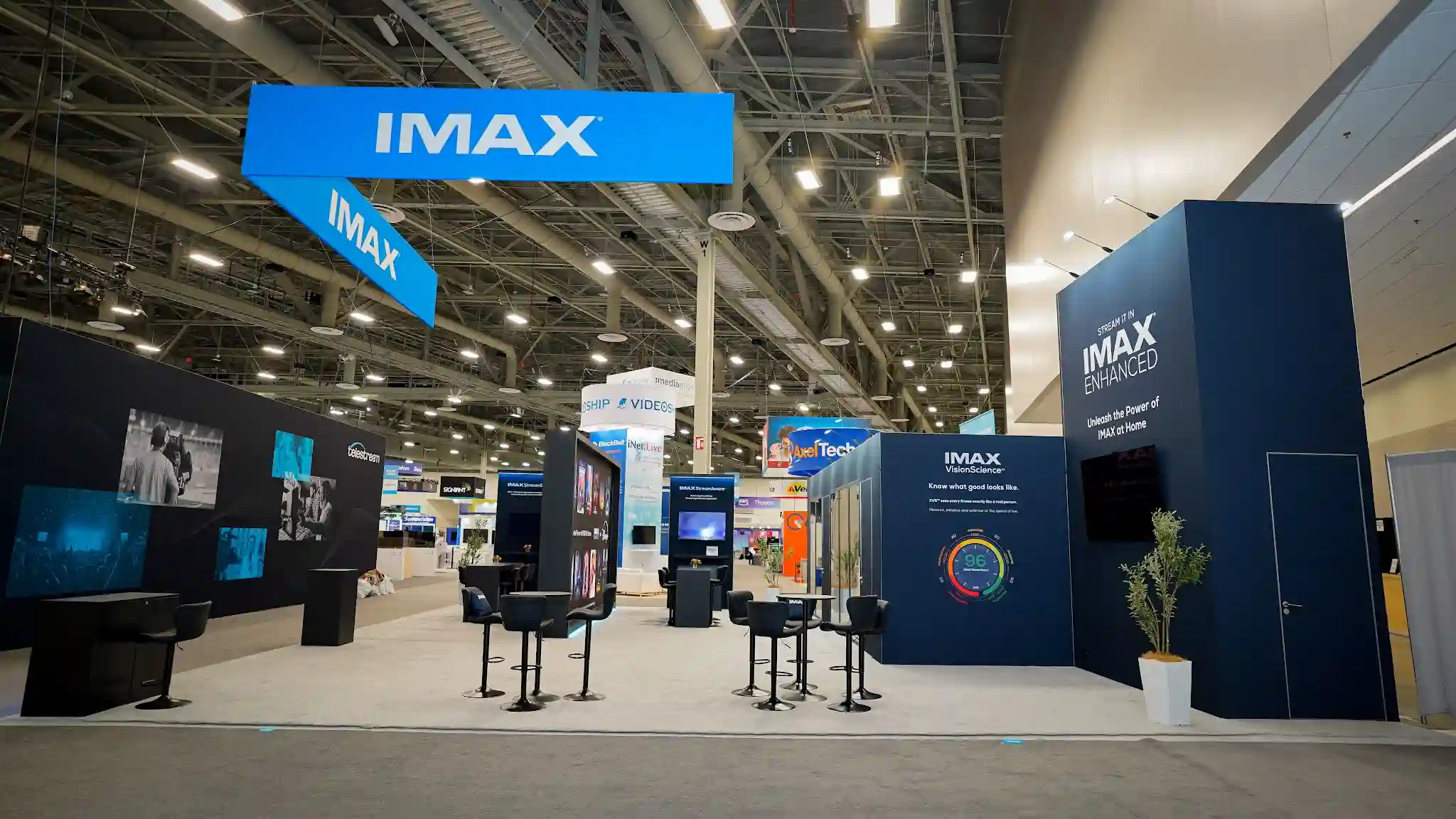 Frontal wide view of IMAX island exhibit showing two-tier hanging sign in navy and cyan, VisionScience wall with Viewer Score 96 graphic, multiple seating areas with branded furniture, dark cinematic walls, and electric blue accent lighting throughout the 2000-square-foot booth at NAB 2025
