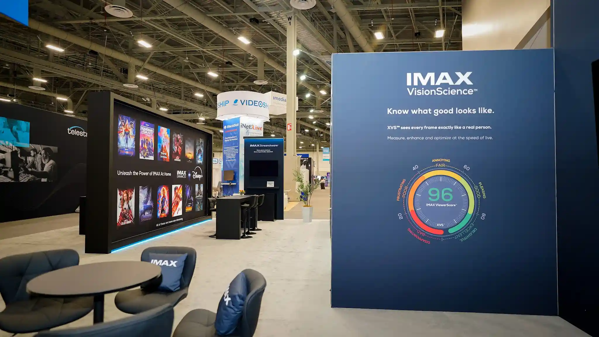 Interior lounge area of IMAX exhibit showing VisionScience interactive display wall with Viewer Score 96 rating graphic against color quality spectrum, movie poster grid showcasing Disney Plus IMAX Enhanced titles, navy blue lounge chairs with branded IMAX pillows, warm ambient lighting, and consultation-ready seating arrangement