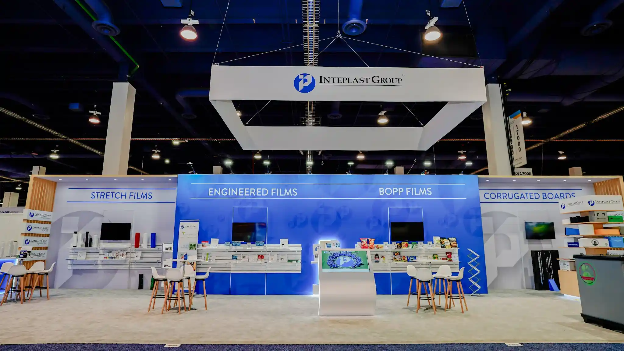 Wide frontal view of Inteplast Group 20x60 island exhibit at PACK EXPO Las Vegas 2025 showing all four product zones — Stretch Films, Engineered Films, BOPP Films, Corrugated Boards — with large rectangular overhead hanging sign bearing Inteplast Group name and blue circular P logo, alternating blue and white color-blocked columns, white slatwall product displays with film rolls, and bar-height meeting tables with wooden-legged stools