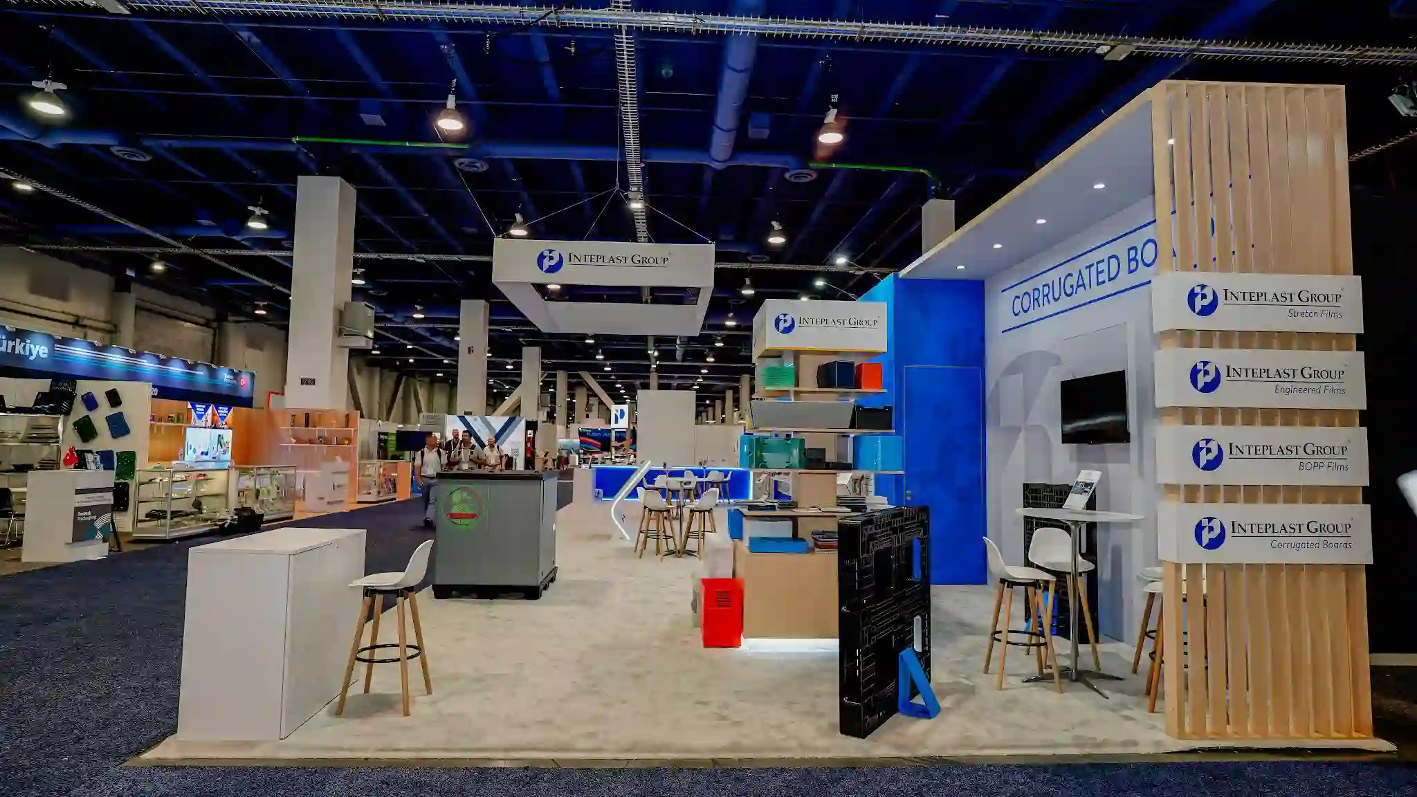 Interior view of Inteplast Group exhibit featuring prominent vertical wood slat wall with integrated division identification panels for Stretch Films, Engineered Films, BOPP Films, and Corrugated Boards, gray industrial machinery display in center foreground, stacked colorful corrugated board product samples, and booth interior with blue and white color-blocked zones in background