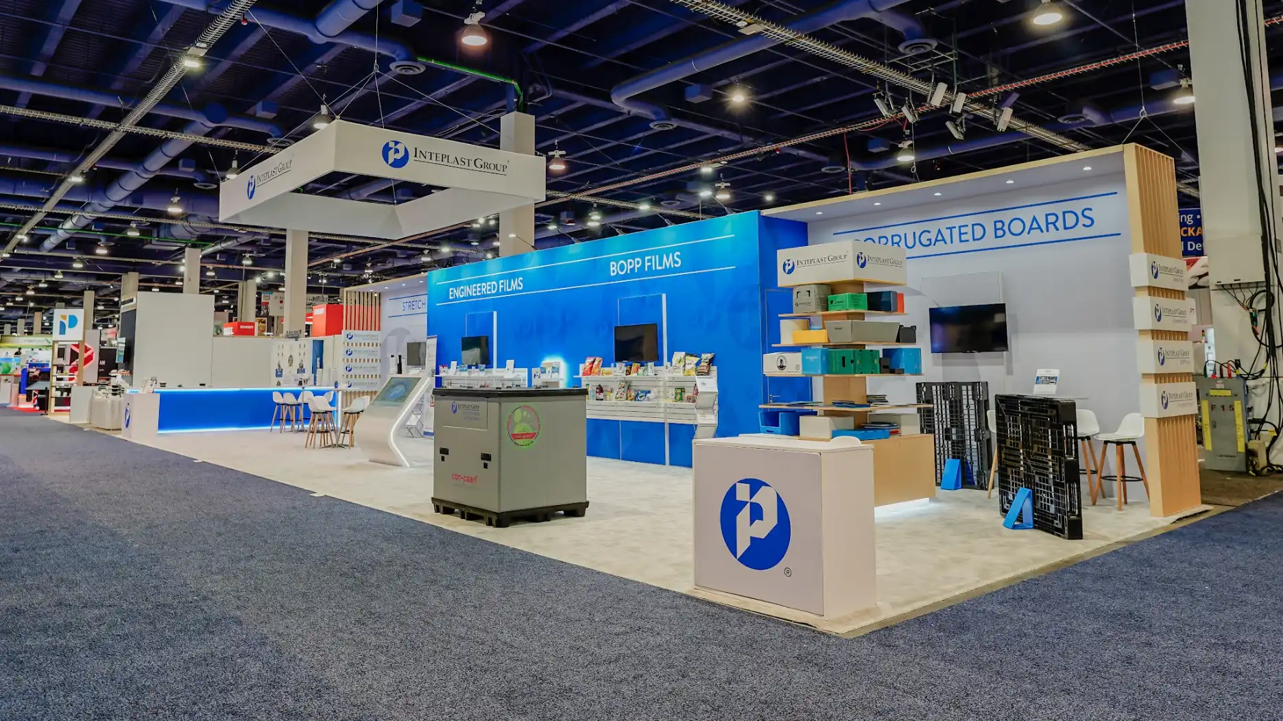 Wide corner view of Inteplast Group island exhibit showing gray industrial machinery display at center, all four product zones visible, stacked corrugated board product samples in yellow, orange, green, blue, and red on right side, full booth scale at PACK EXPO Las Vegas 2025