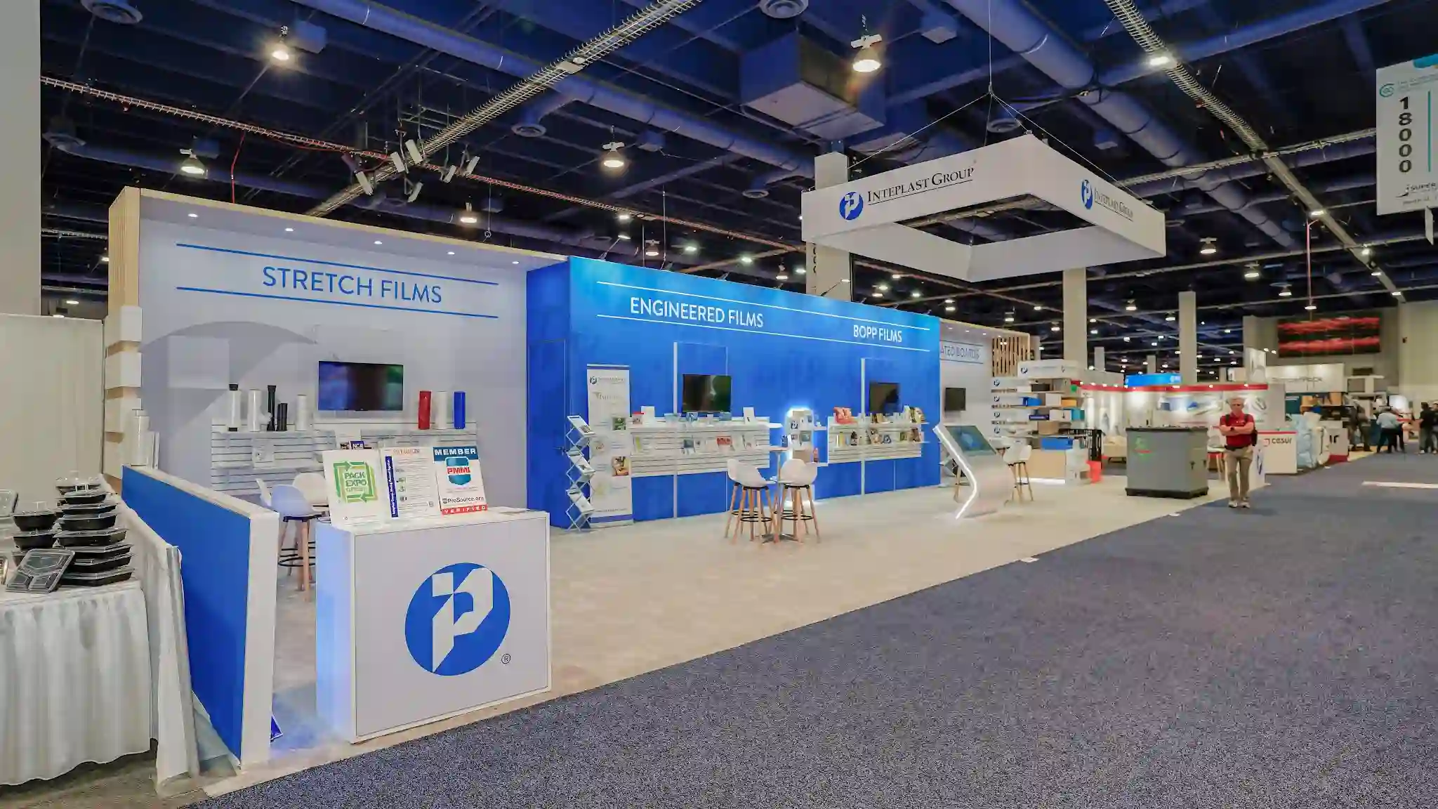 Angled corner view of Inteplast Group island exhibit at PACK EXPO Las Vegas showing white reception counter with large Inteplast Group logo at corner entrance, FRESH LOCK branded product displays, stacked black product cases, bar stools and meeting tables in background, providing depth perspective across the 1,200-square-foot booth footprint