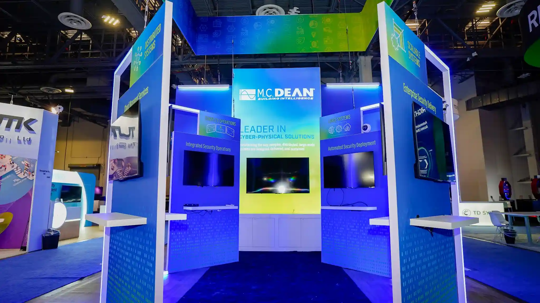 M.C. Dean central kiosk hub close-up showing Leader in Cyber-Physical Solutions headline, redefining the way complex distributed large-scale systems are designed delivered and sustained tagline, Complex Operations Integrated Security Operations panel, Large Systems Automated Security Deployment panel, widescreen monitors with digital lens flare graphics, blue LED base glow, and white interaction counters