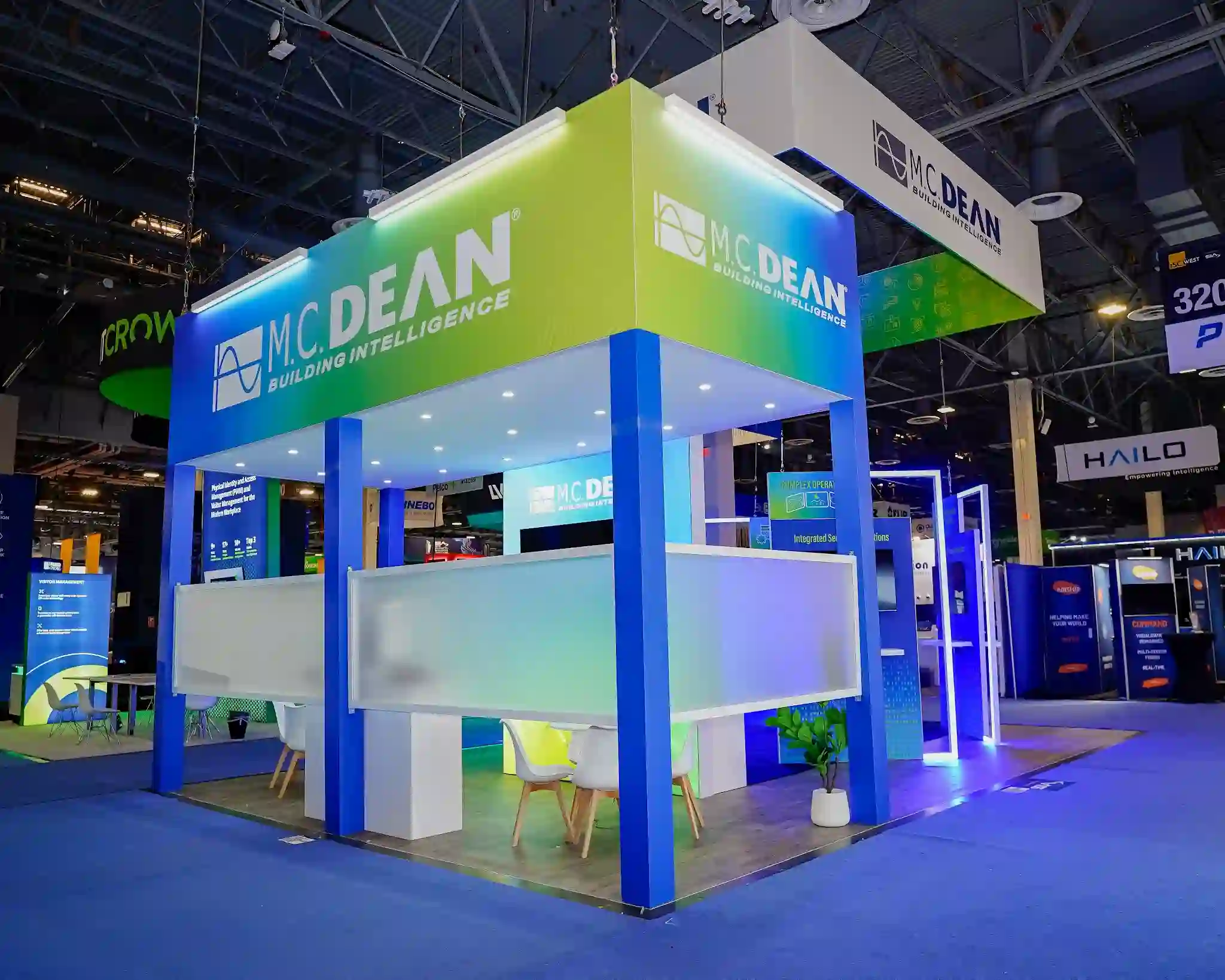 M.C. Dean semi-enclosed meeting lounge with thick blue vertical pillars, lime-green branded canopy reading M.C. Dean Building Intelligence, frosted privacy panels, warm wood-style flooring, white Eames-style chairs, circular tables, potted plant, and views through to central kiosk demos in background